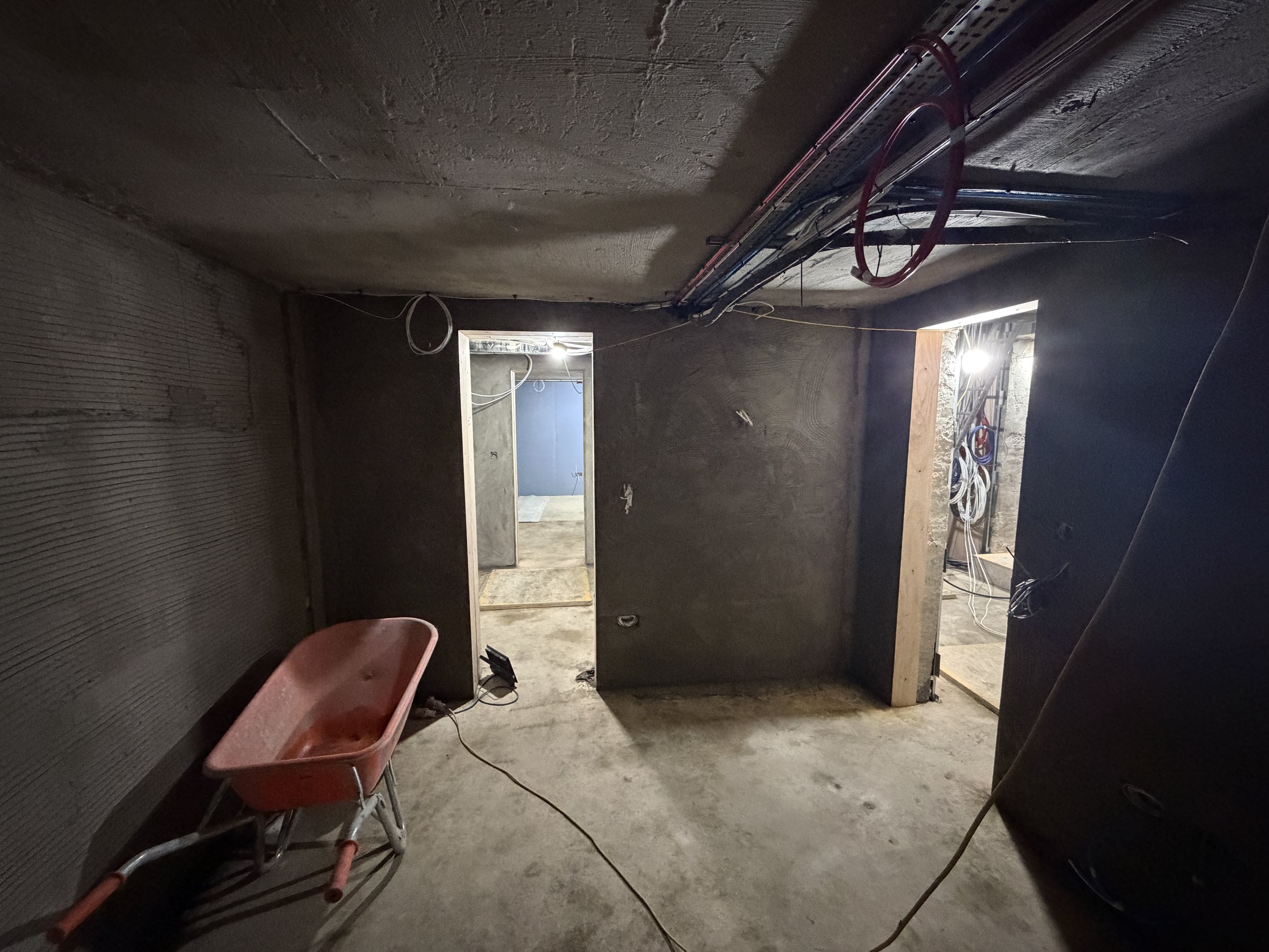 Inside a building under construction with unfinished walls and exposed wiring. A red plastic wheelbarrow is in the foreground, and there's a doorway leading to another room with a small window, partially illuminated lights.