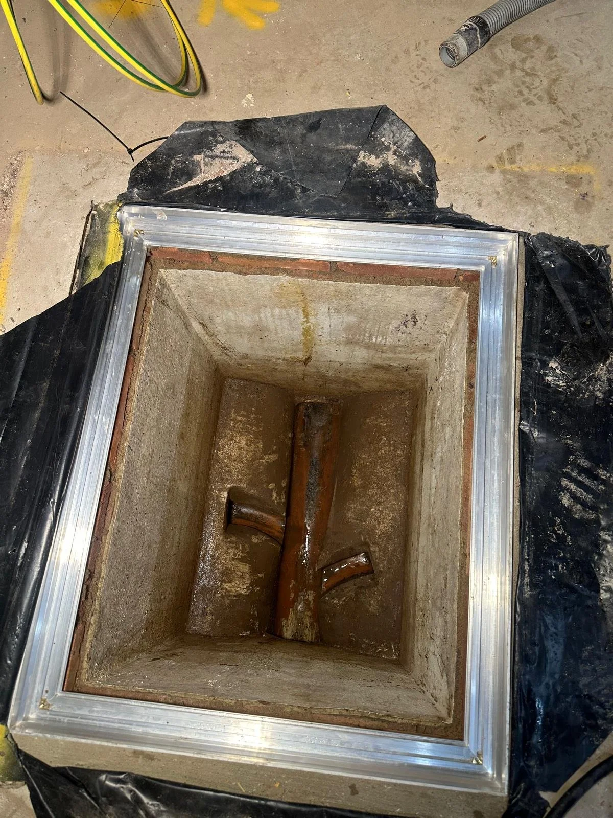 A rectangular underground utility access box with a concrete interior, containing rusted pipes and a metal pipe elbow, surrounded by black plastic sheeting, on a concrete floor with yellow markings.
