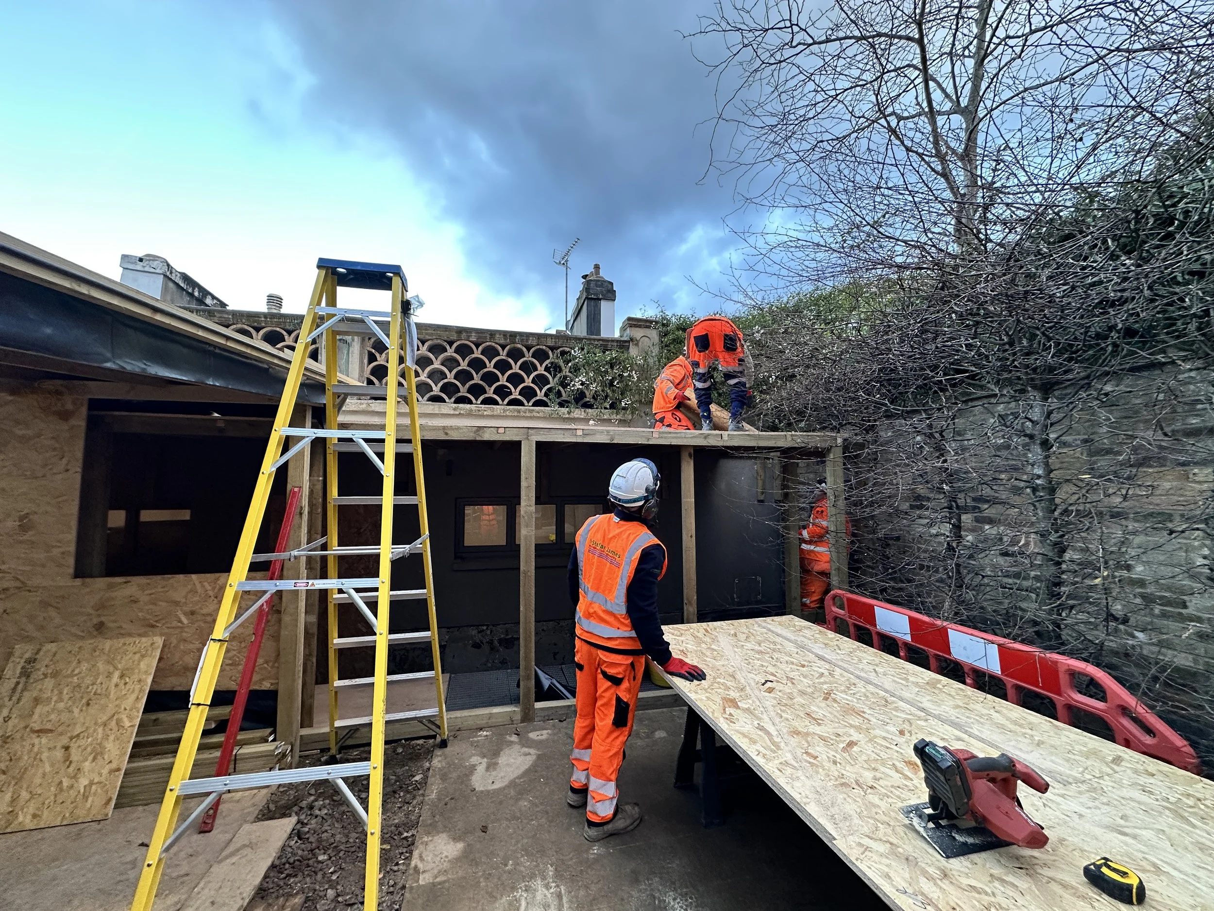 Construction workers in high visibility clothing working on a building project outdoors during daytime, with trees and cloudy sky in the background.