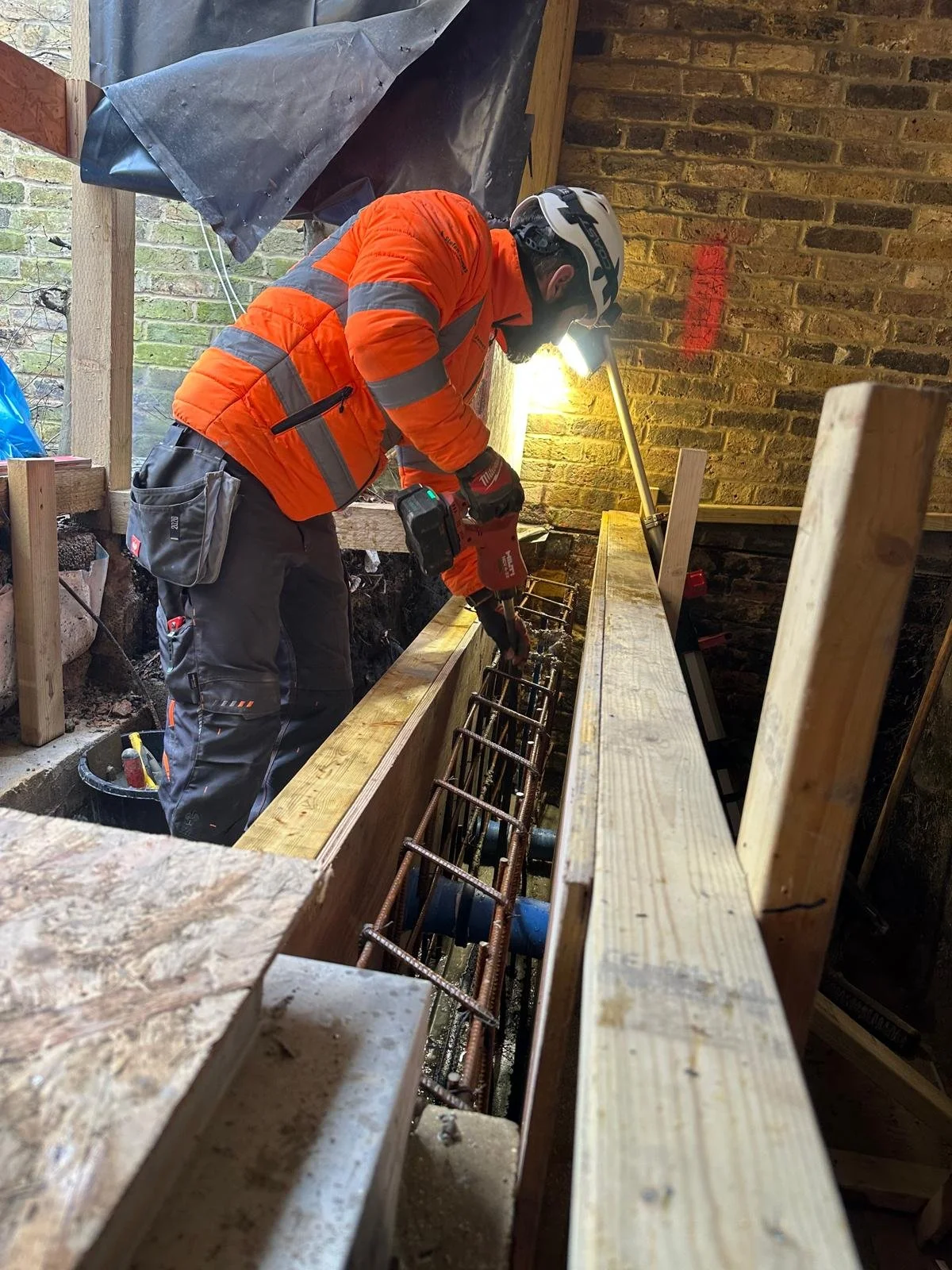 A construction worker wearing an orange jacket, gray pants, and a white helmet is working on a wooden frame with rebar reinforcement inside a building with a brick wall. The worker is using a power drill, and there is a bright work light illuminating the area.
