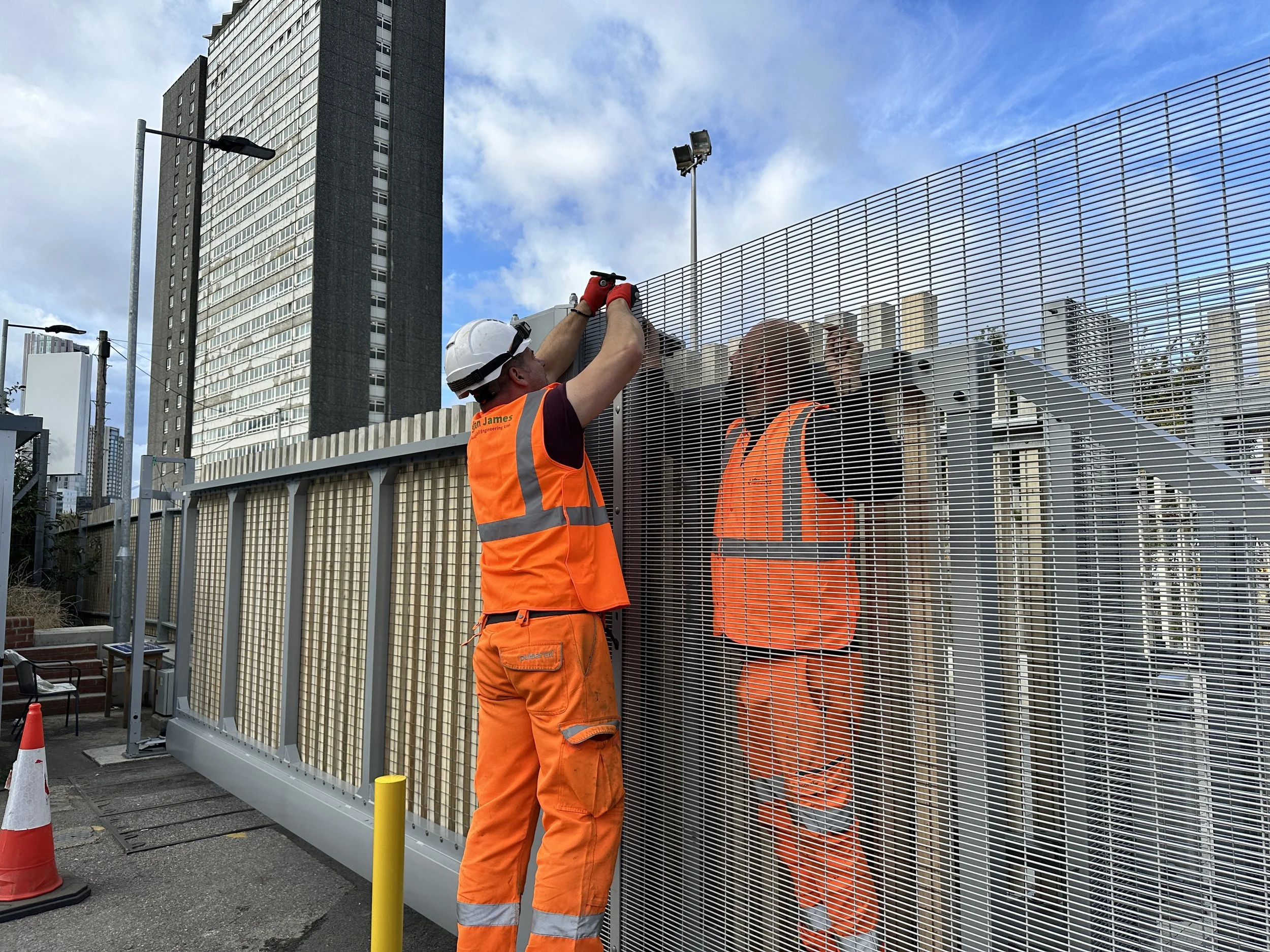 Two workers in orange safety vests and helmets installing or repairing a metal fence on a city street with high-rise buildings in the background.