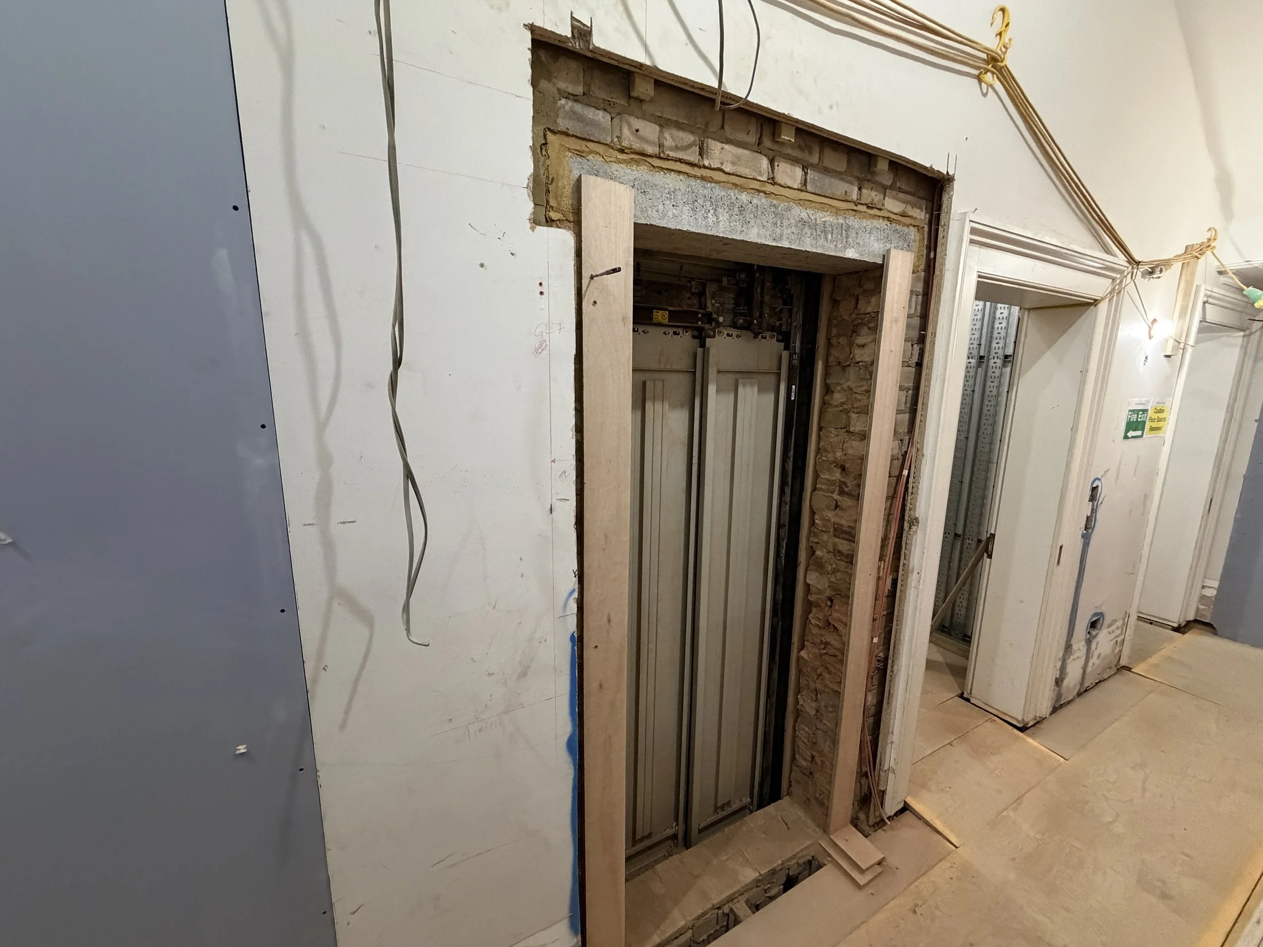 The image shows an indoor construction site with a partially removed wall, revealing brick and concrete behind it. There are wooden and metal framing components, exposed wiring, and adjacent areas under construction, including a door and fire safety signs.