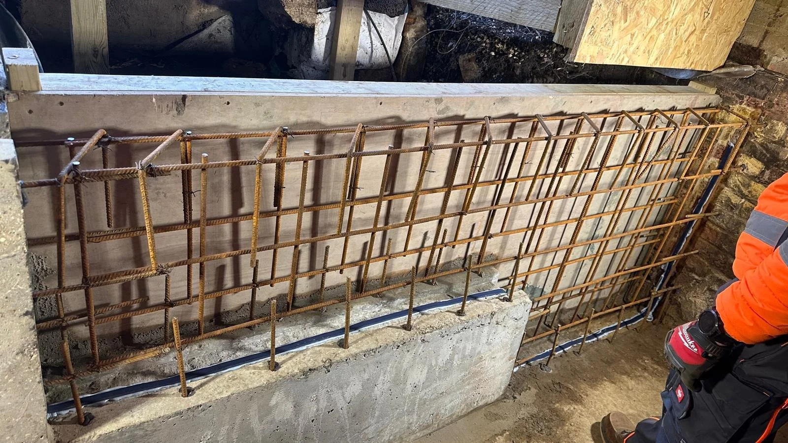 Steel rebar grid installed over a concrete foundation, part of a construction site, with a worker in safety gear on the right.