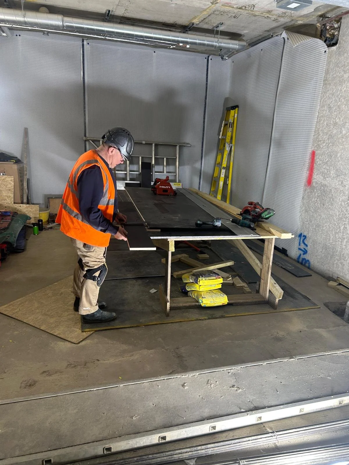 A construction worker wearing a black helmet, orange safety vest, and tan work pants stands at a workbench in an unfinished building. The workspace includes tools, materials, and a ladder in the background. The worker appears focused on a project at the table.