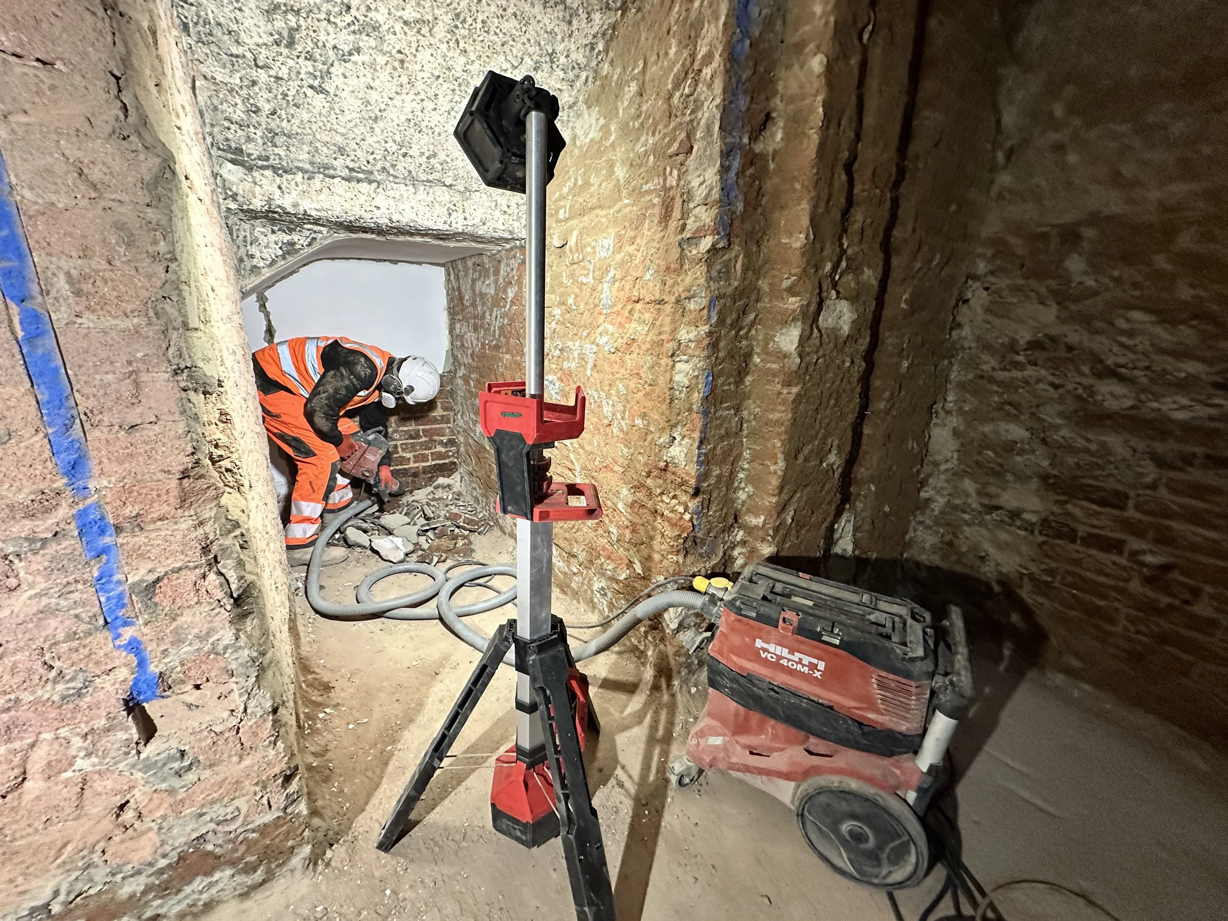 Construction worker in safety gear working in a brick underground tunnel. Construction equipment, including a laser level and vacuum, is in the foreground.