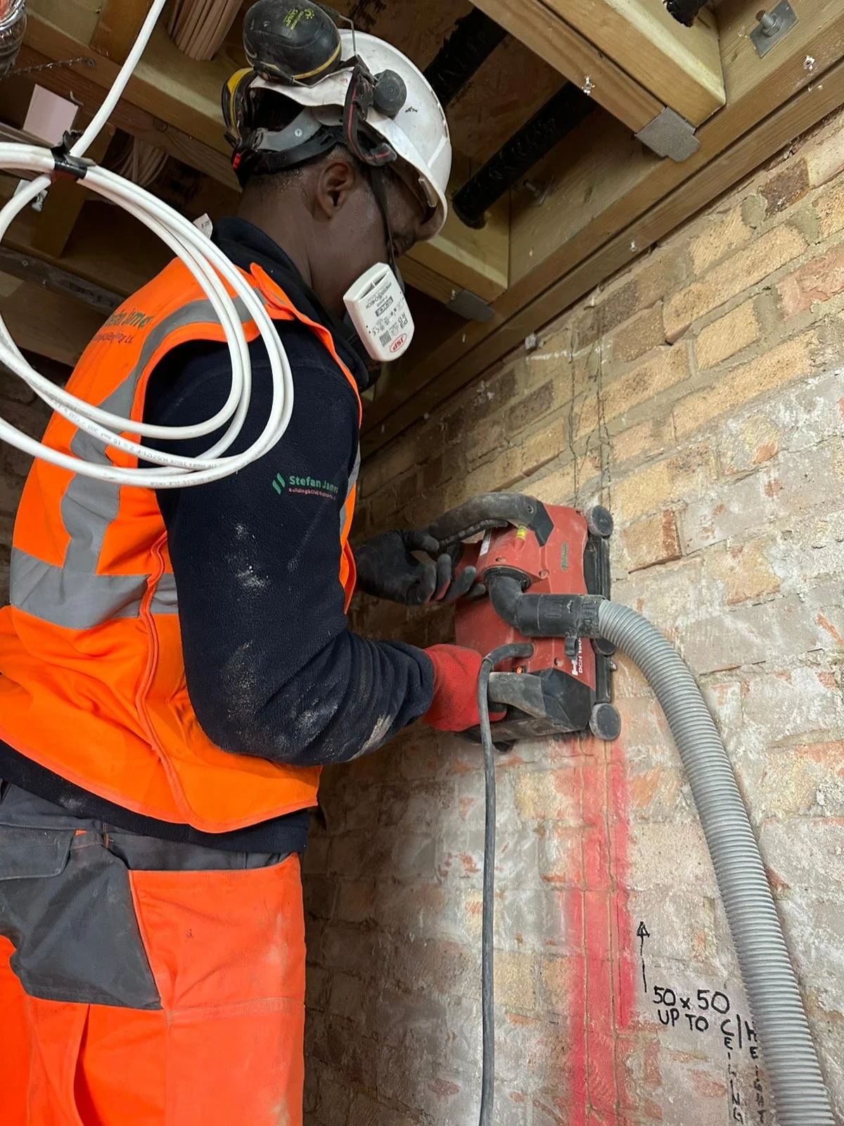 A construction worker in an orange safety vest, black shirt, and safety gear, including a helmet and face mask, is using a power drill on a brick wall in a construction site. There are construction notes and measurements written on the wall.