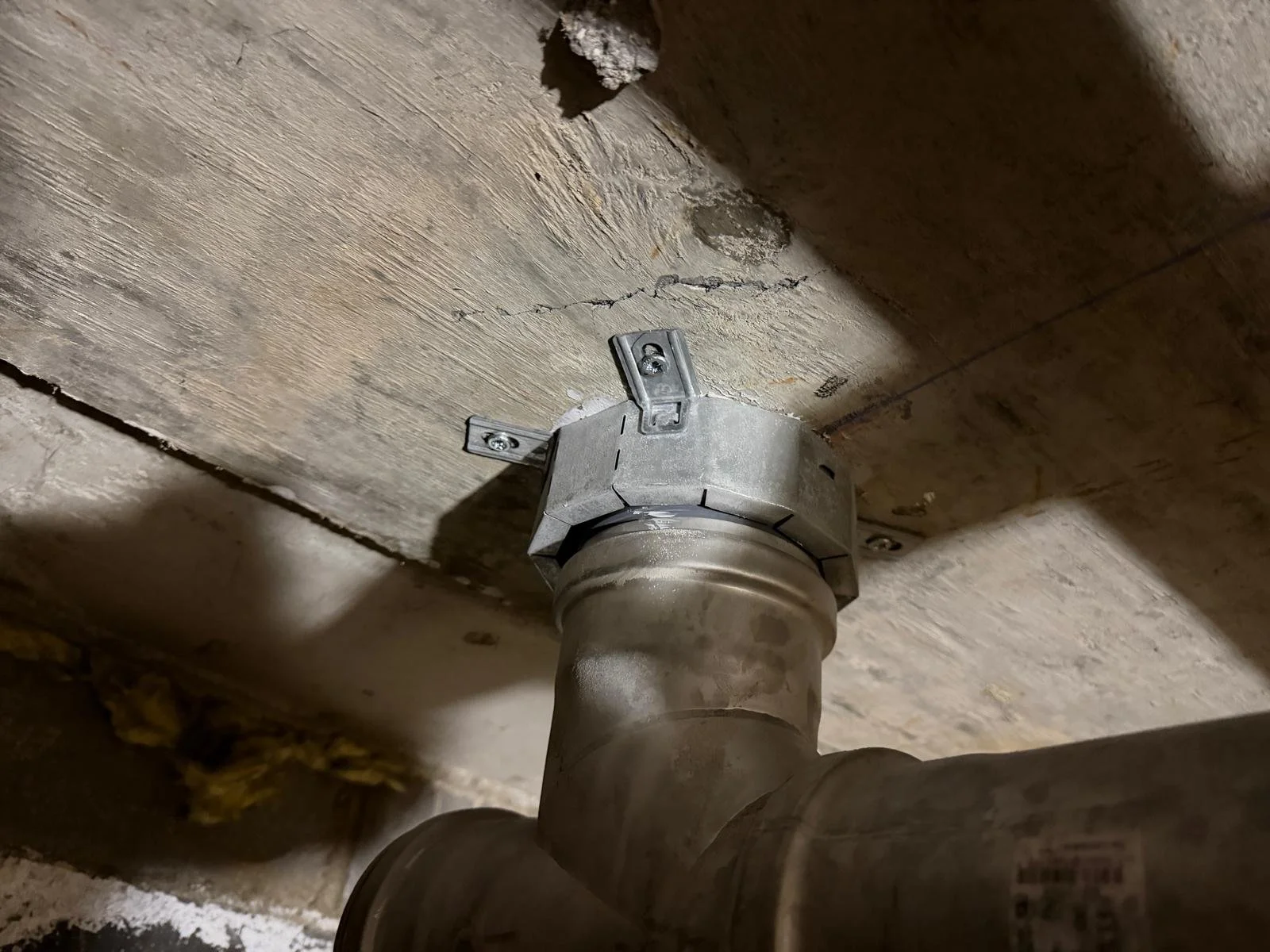 Close-up of a metal pipe connector attached to a plumbing pipe underneath a wooden floor with visible cracks.