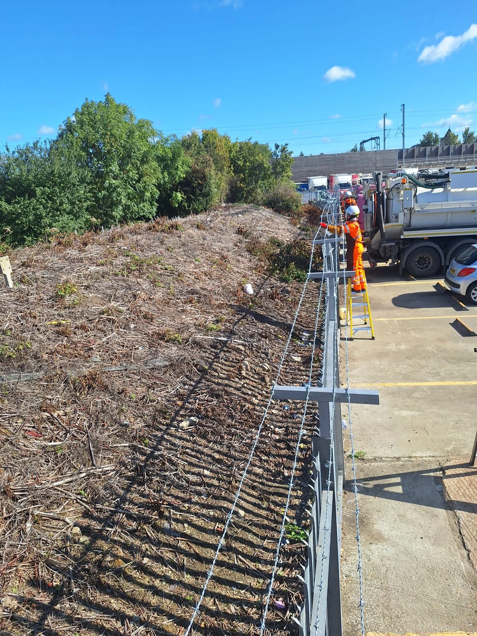 Construction worker in orange safety gear repairing a security fence along a dirt hillside with trees in the background and a parking lot with trucks and cars nearby under a clear blue sky.