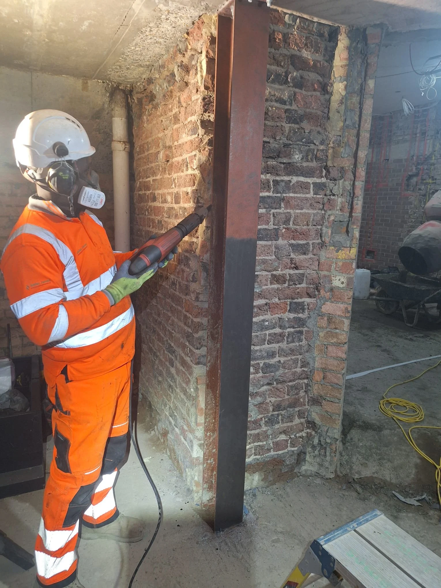 Worker in orange safety uniform and helmet operating a power tool on a brick wall at construction site.
