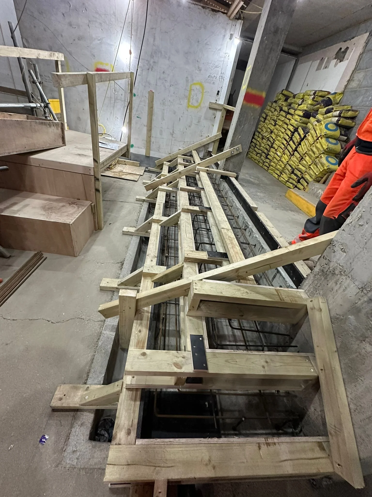 Construction site with wooden frame for a structure, stacks of bags on the side, and a person wearing orange work clothes.