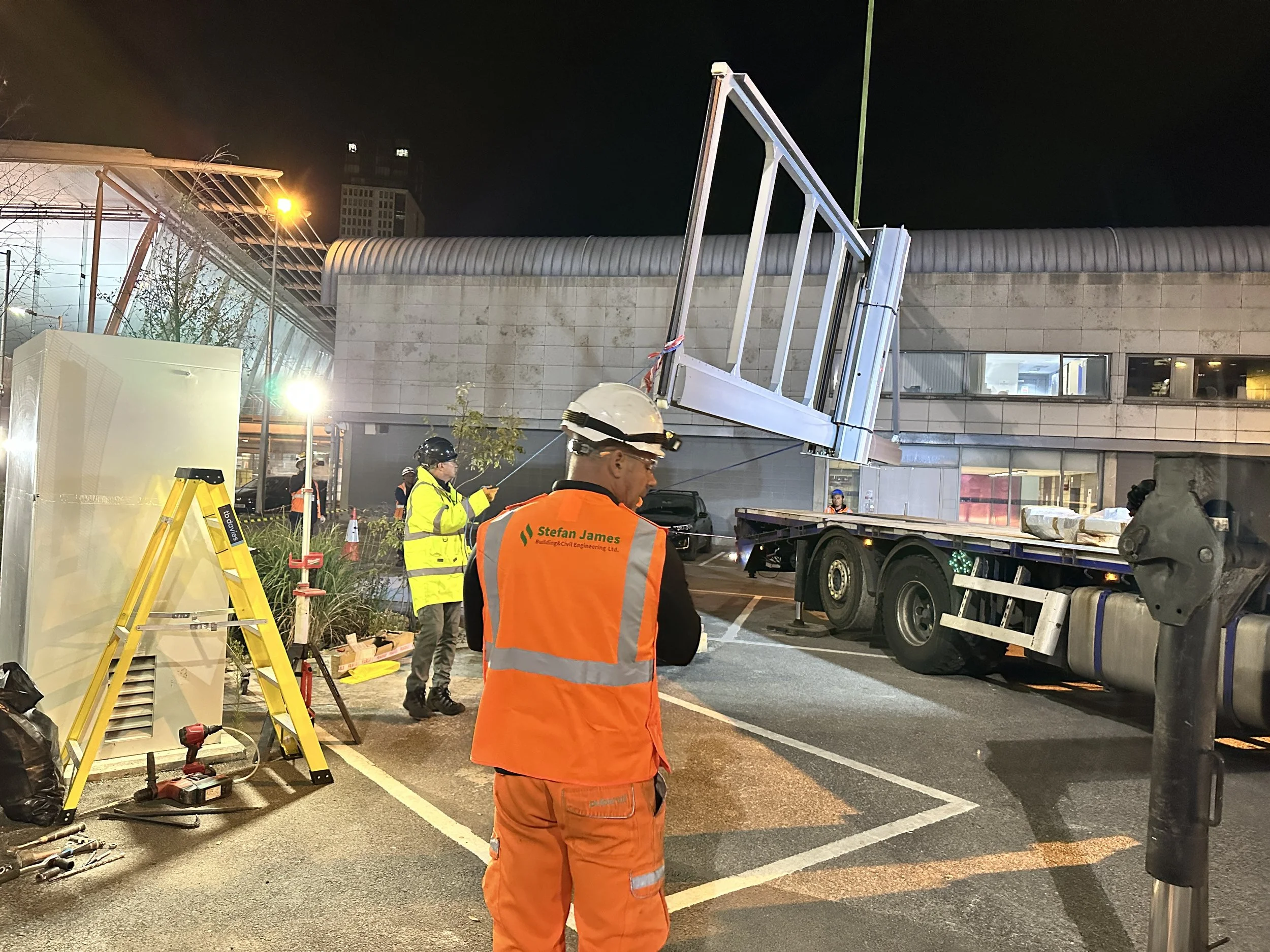 Construction workers in high-visibility clothing install a large billboard or sign on a building at night. One worker is holding a tool near the billboard, while others are nearby, with a truck parked alongside.