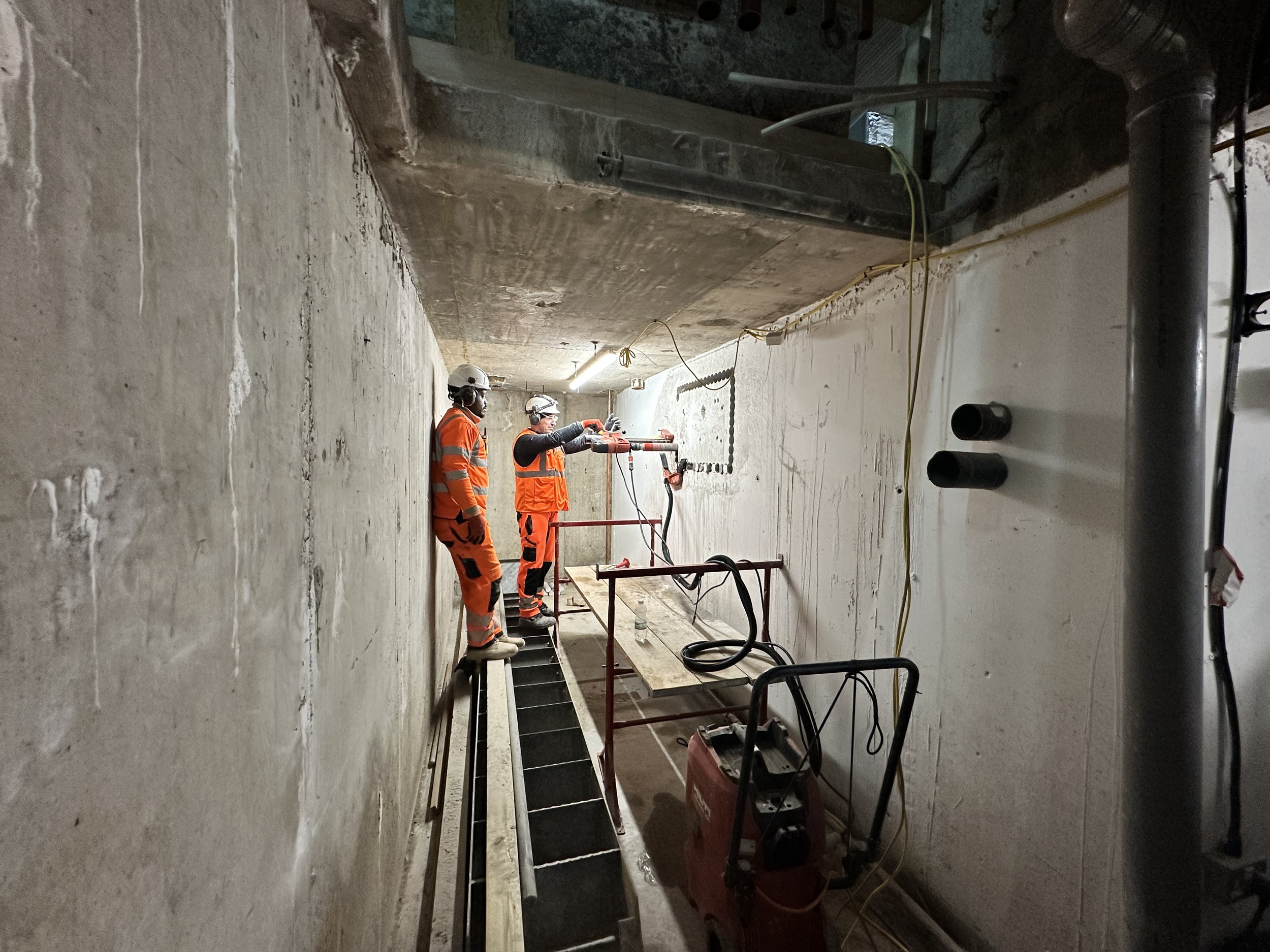 Construction workers wearing safety helmets and orange safety gear working inside a concrete structure, using tools and equipment for building or renovation.