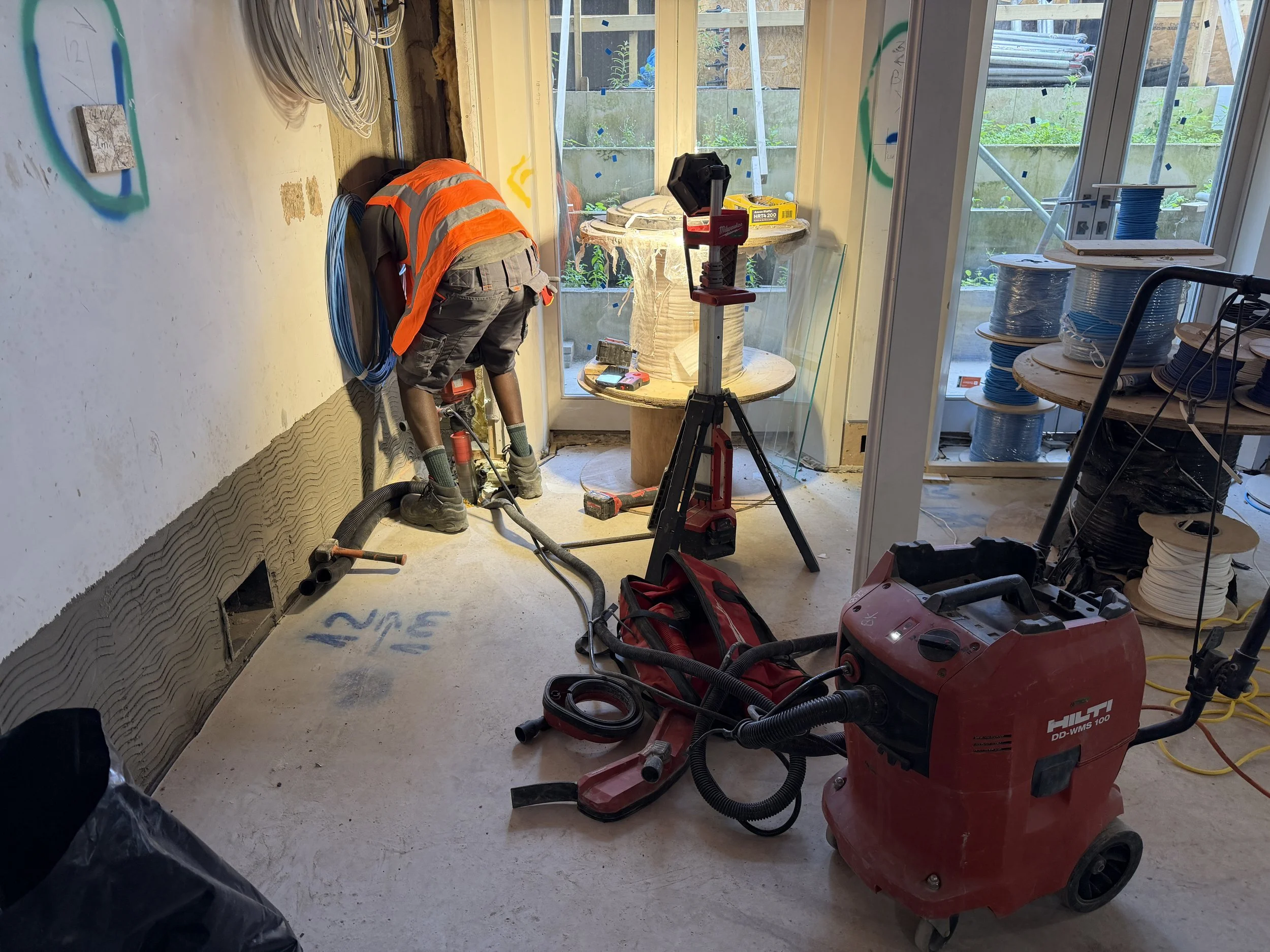 A construction worker in a safety vest is kneeling on the floor, working on electrical wiring inside a room under renovation. The room has large glass doors leading outside, and construction tools and supplies are scattered around.