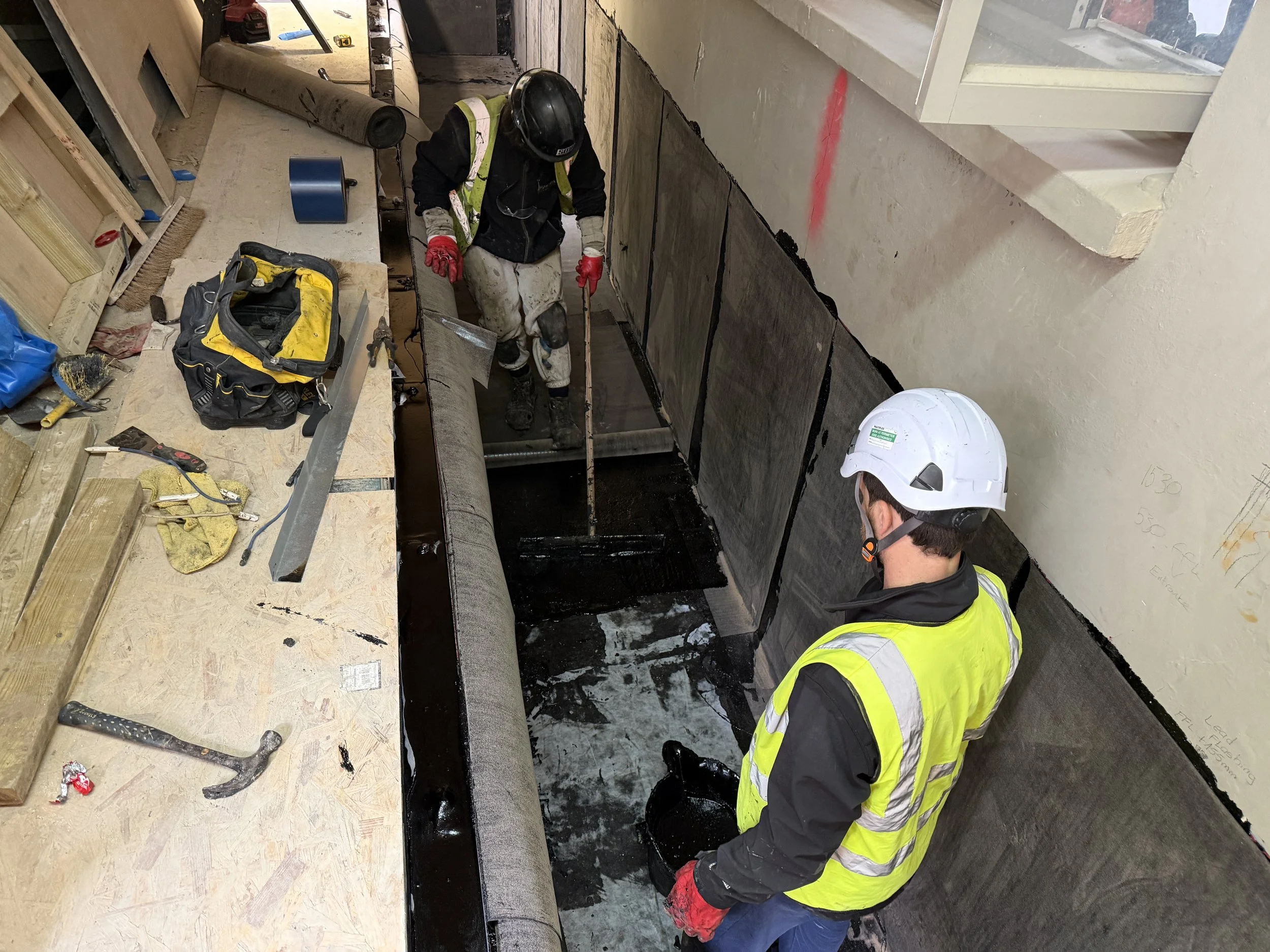 Two construction workers wearing yellow safety vests and helmets are working on a building interior. They are installing or repairing a black waterproof membrane along a wall with a large window, with tools and supplies scattered around.
