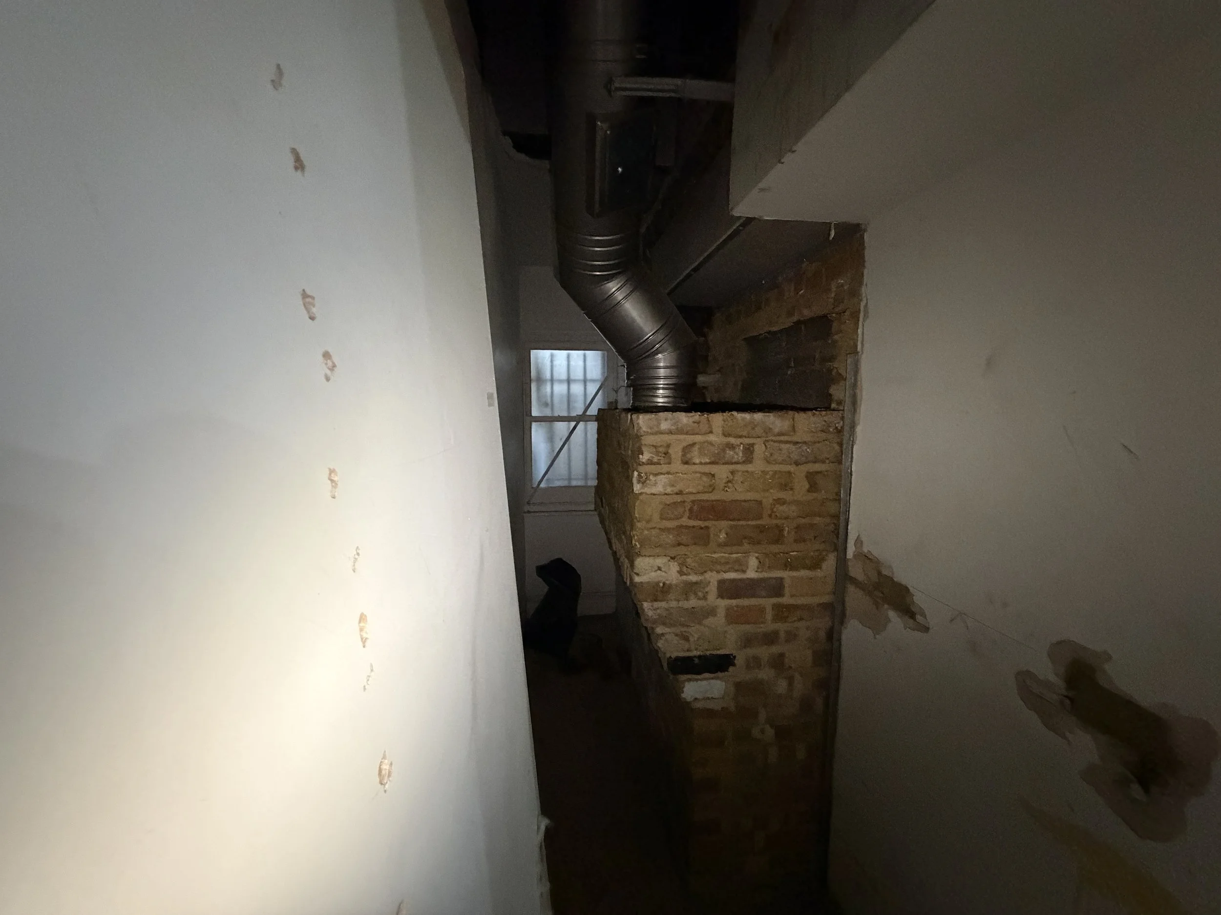 Construction site showing an unfinished brick wall, exposed ductwork, and an opening to another room with visible window panels.