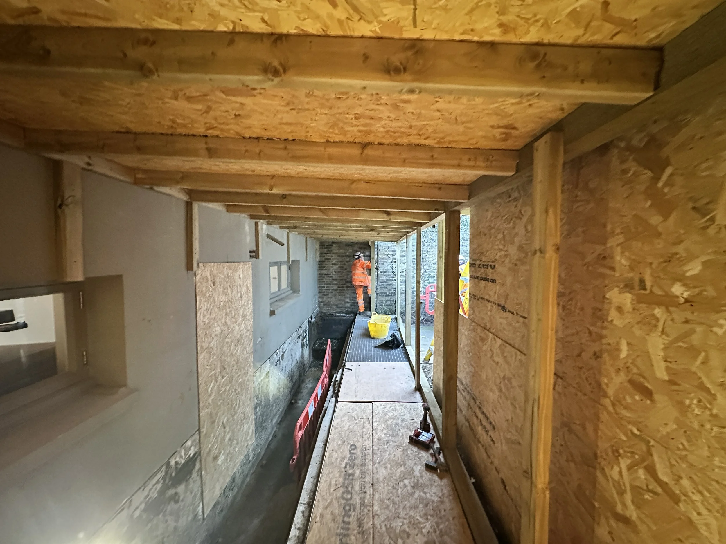 Construction workers in orange safety vests working on framing a building with wooden studs and plywood.