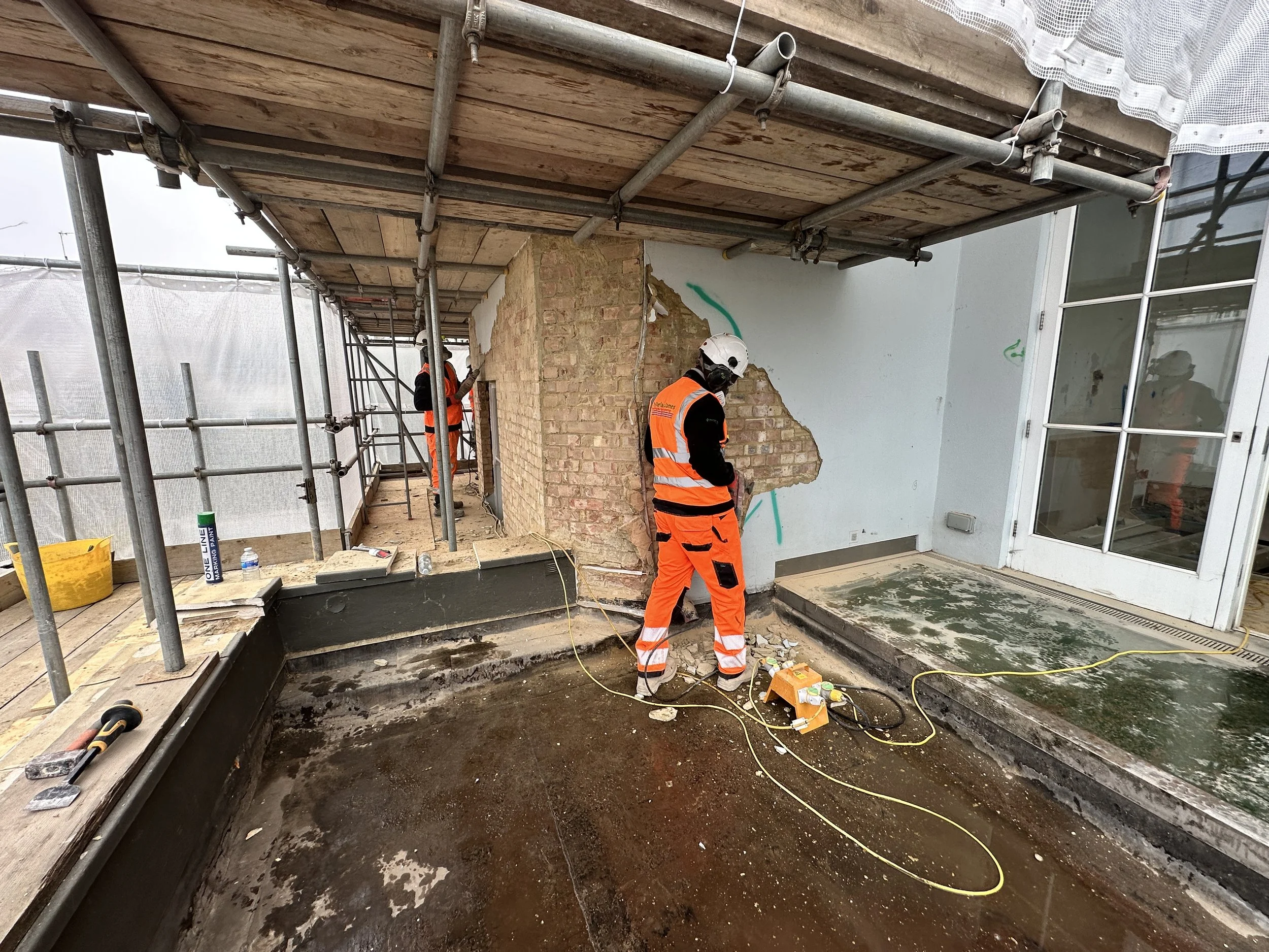 Construction workers in orange safety vests and helmets working on building renovation, with scaffolding, a brick wall, and a concrete foundation.