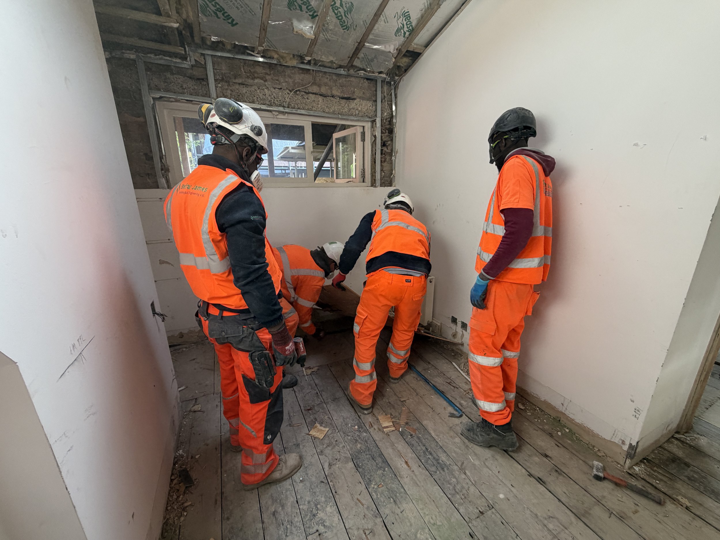 Construction workers wearing safety gear working inside a partially demolished room, with exposed ceiling and debris on the floor.