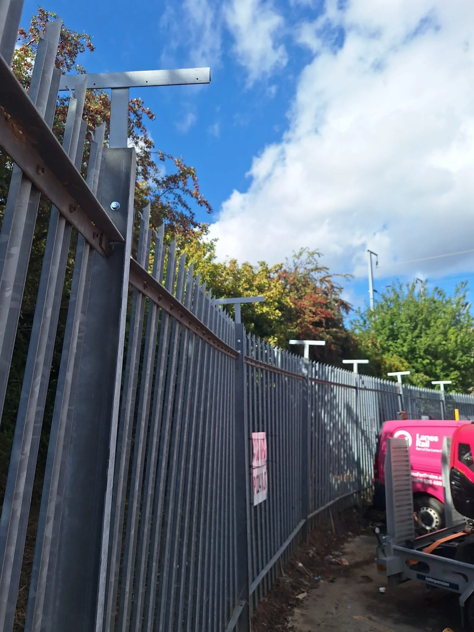 A metal fence with vertical bars and attached horizontal supports runs along a dirt pathway. In the background, trees with green and reddish leaves, blue sky with some white clouds, and several white streetlights are visible. On the right, there is a pink utility vehicle parked, and some construction or maintenance equipment, including a trailer, is also present.
