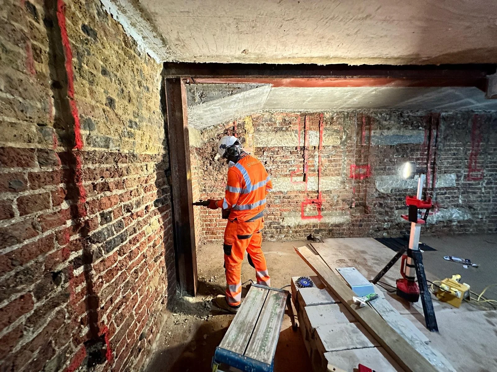 A construction worker wearing an orange safety uniform and helmet is working on a brick wall inside an unfinished building, with tools and construction materials around.