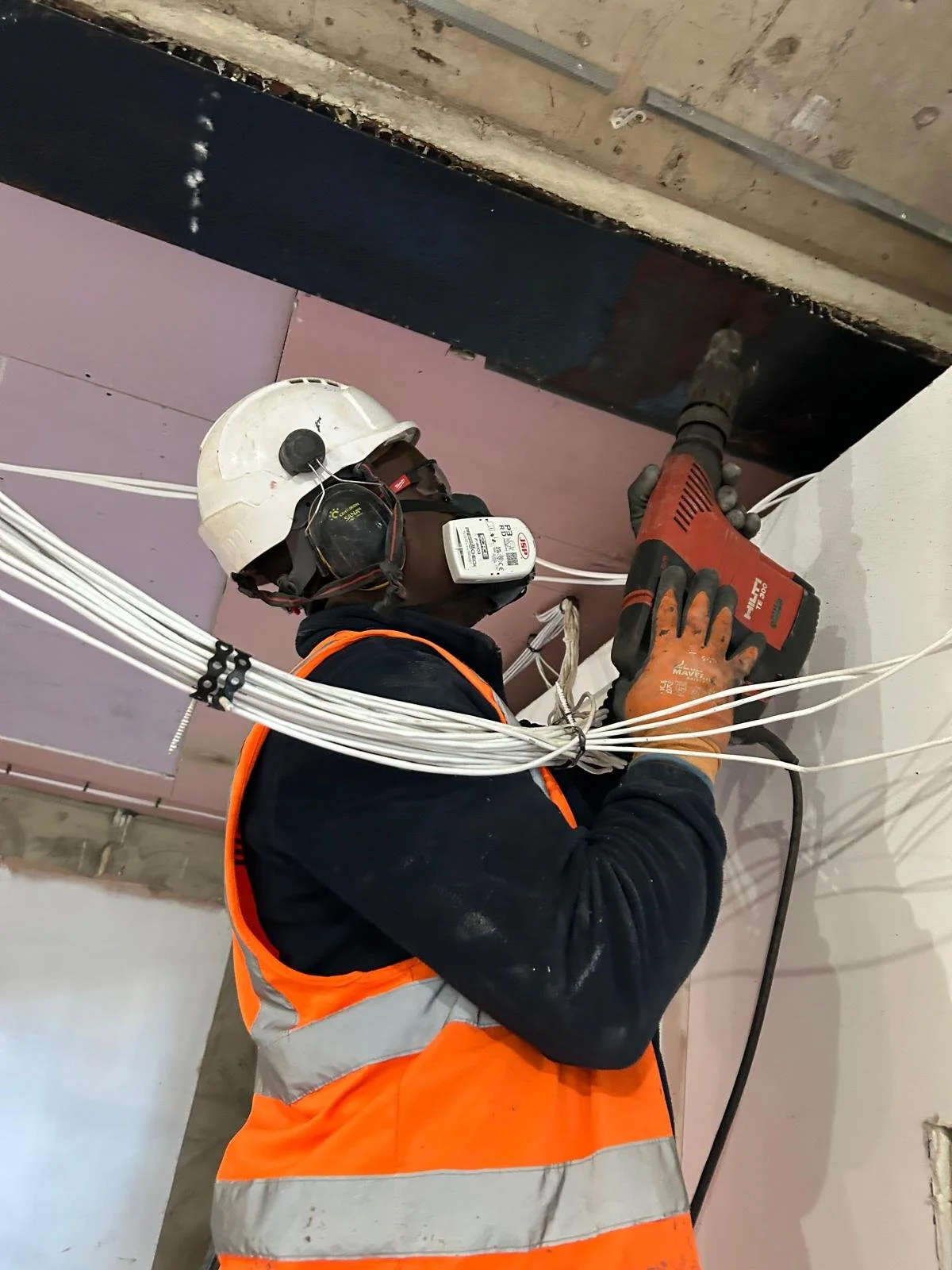 Construction worker wearing a safety helmet, high-visibility vest, and gloves is using a power drill to drill into the wall while surrounded by electrical wires.