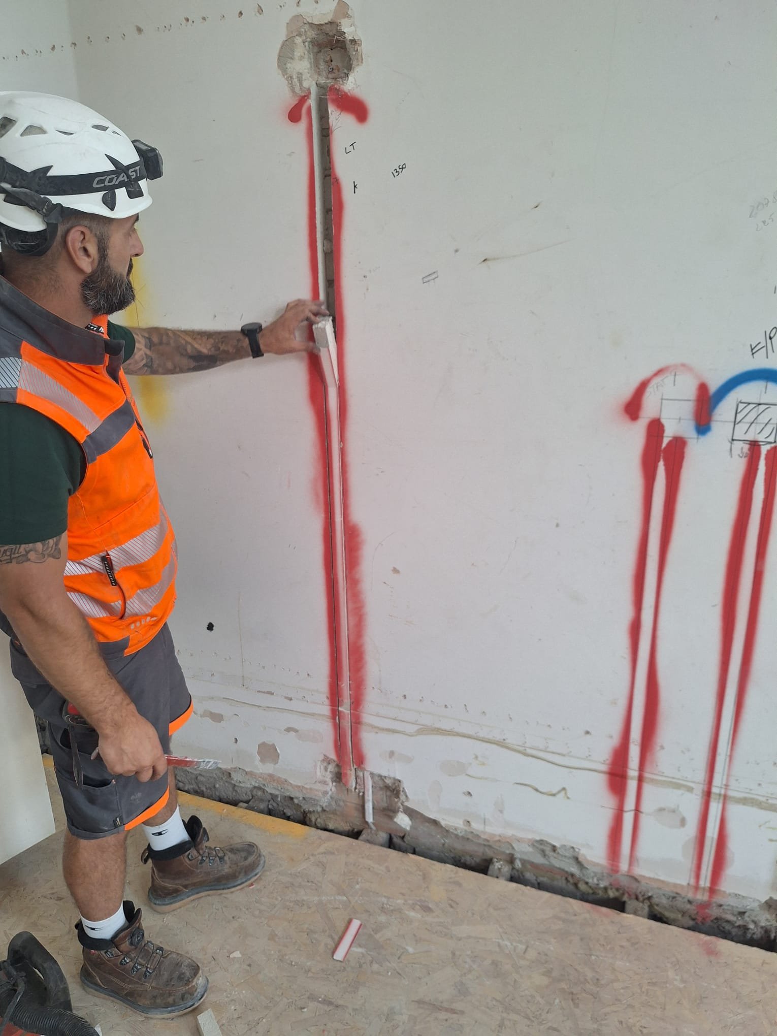 A construction worker wearing a white helmet, orange safety vest, and brown boots standing next to a wall with a hole, exposing pipes and structural elements at the bottom. The worker is pointing at the wall with his right hand.