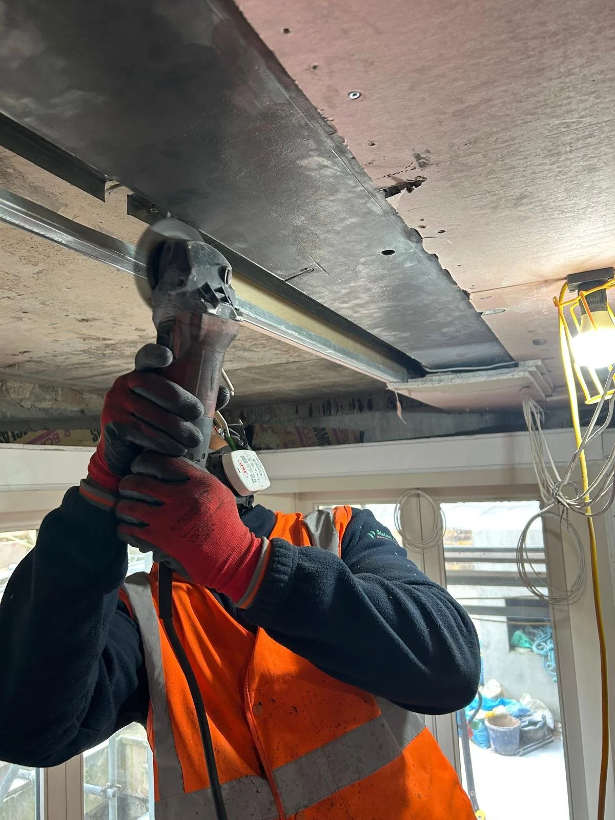A construction worker wearing a safety vest and gloves is using a power drill to secure a metal frame to the ceiling inside a building under construction.