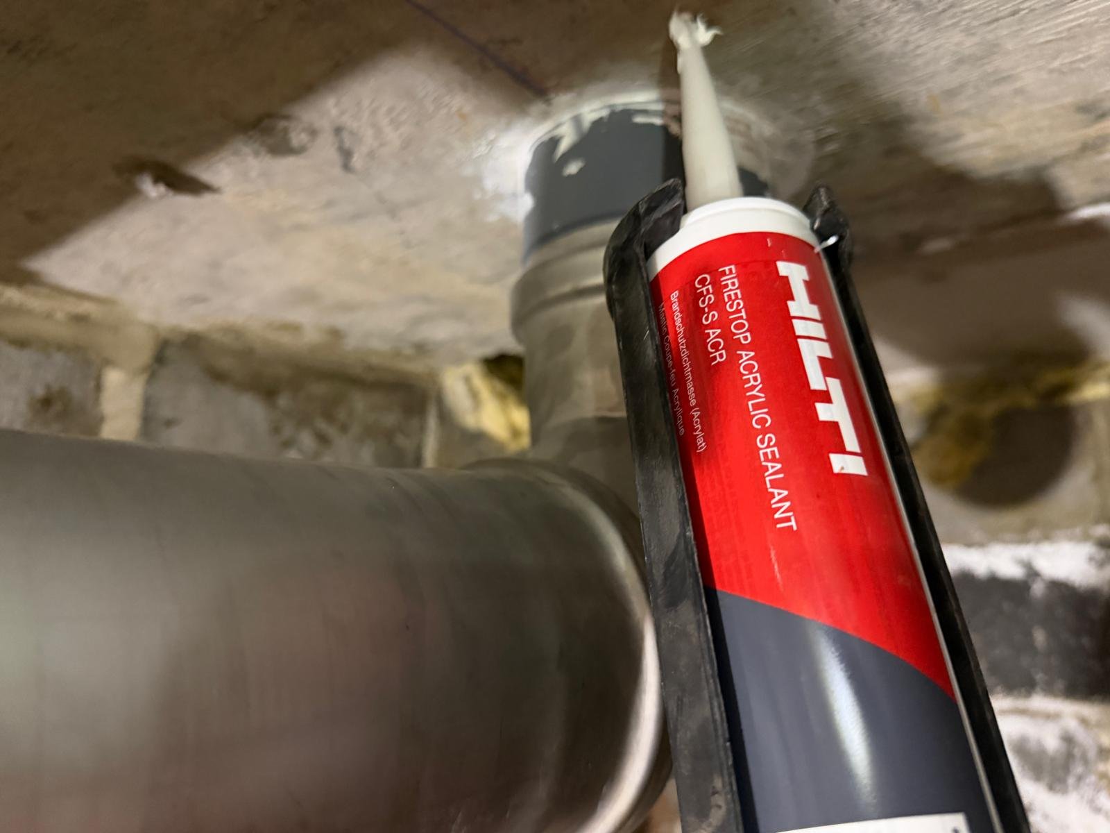 A caulking gun tube labeled 'Hilti Firestop Acrylic Sealant' is positioned against a wall, with a partially applied white sealant in an opening in the wall.