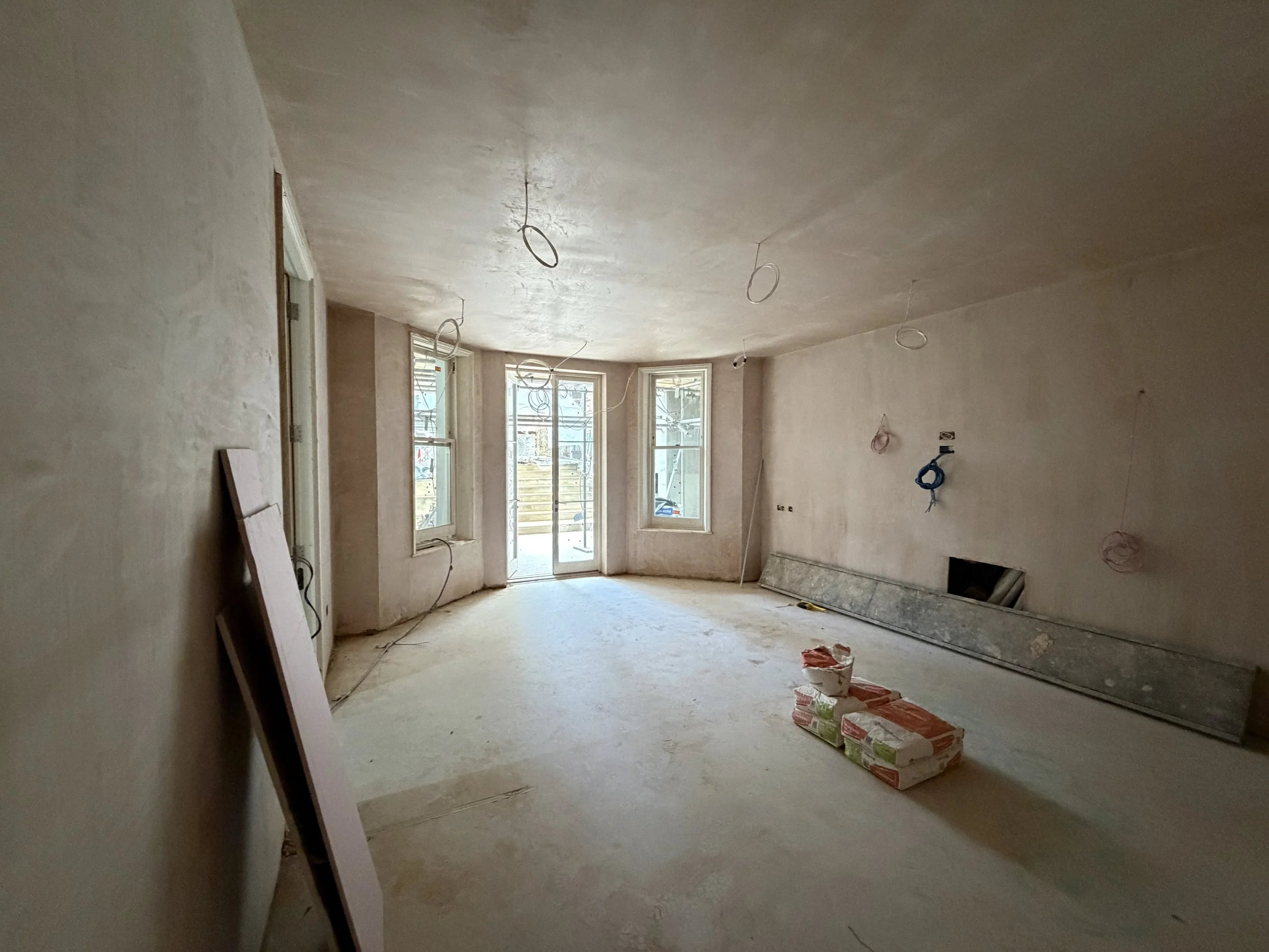 Interior of a house under construction with unfinished walls, exposed wiring, and construction supplies on the floor, including bags and a large panel leaning against the wall, with sunlight coming through the windows.