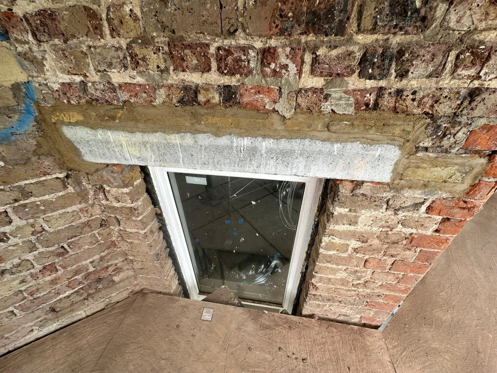 A brick wall with a small rectangular window installed in it, with an exposed brick finish and construction materials around the window.