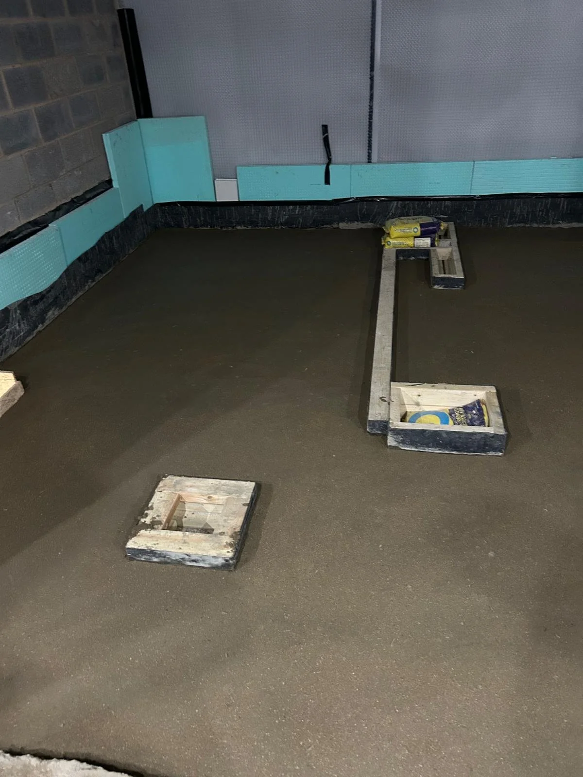 Concrete floor with wooden frame and some tools or materials for construction, with insulation boards on the wall and a black plastic sheet along the edge.