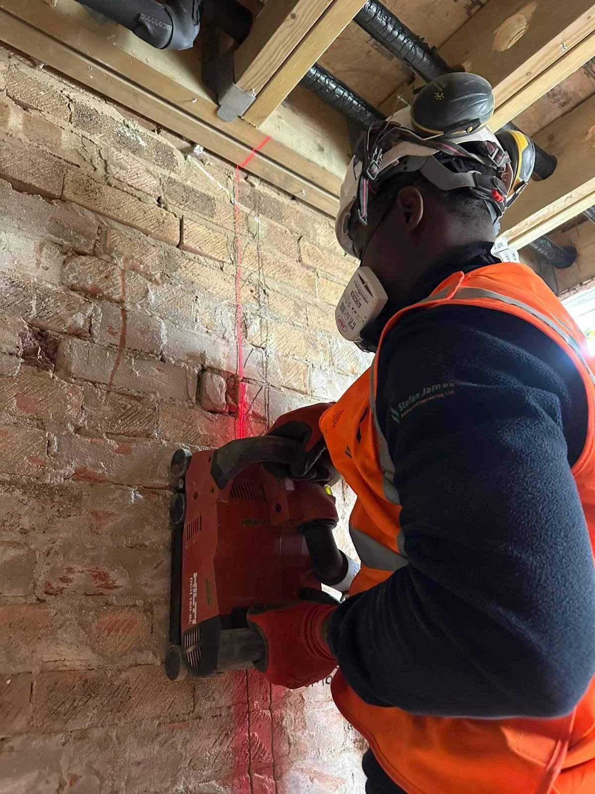A construction worker wearing safety gear using a power drill to make a hole in a brick wall.