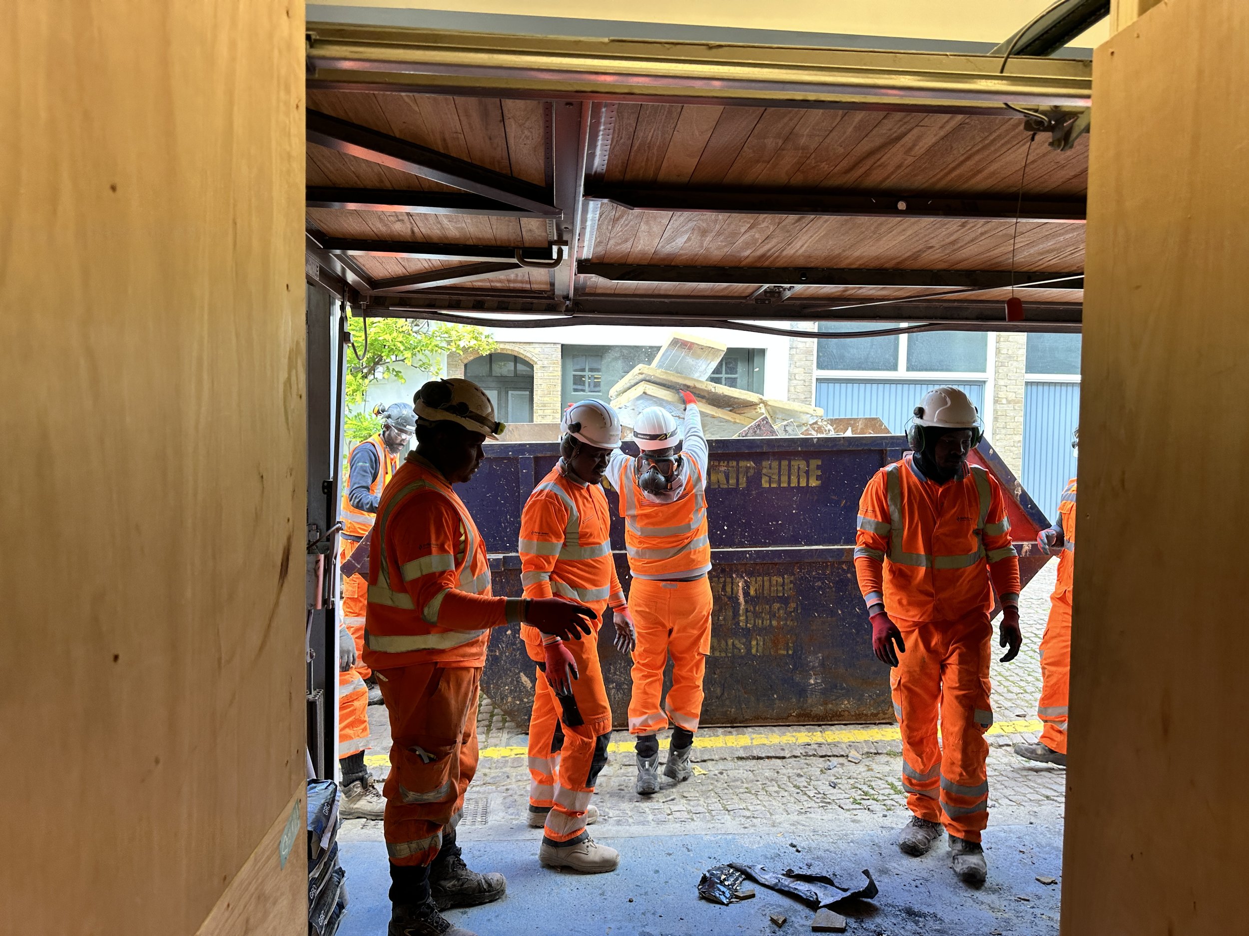 Construction workers wearing orange safety uniforms and white helmets, working outdoors near a building, with some debris and equipment visible.