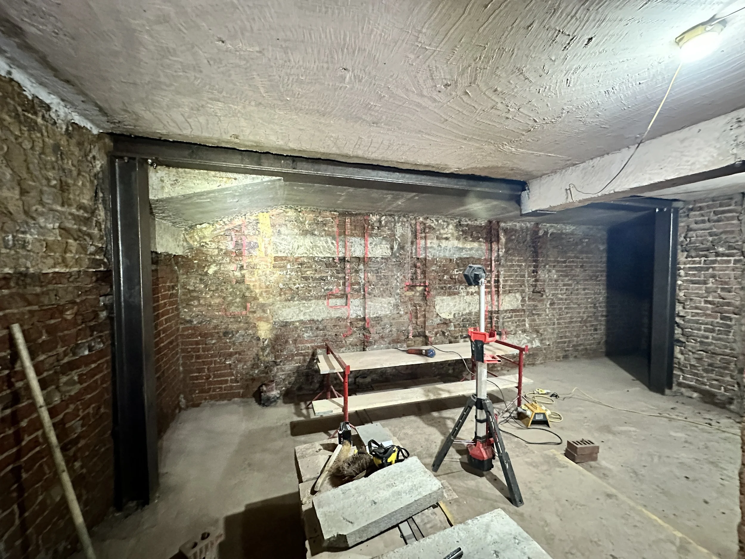 Construction site with brick walls, metal support beams, and tools, including a drill and a workbench with bricks, on an unfinished floor.