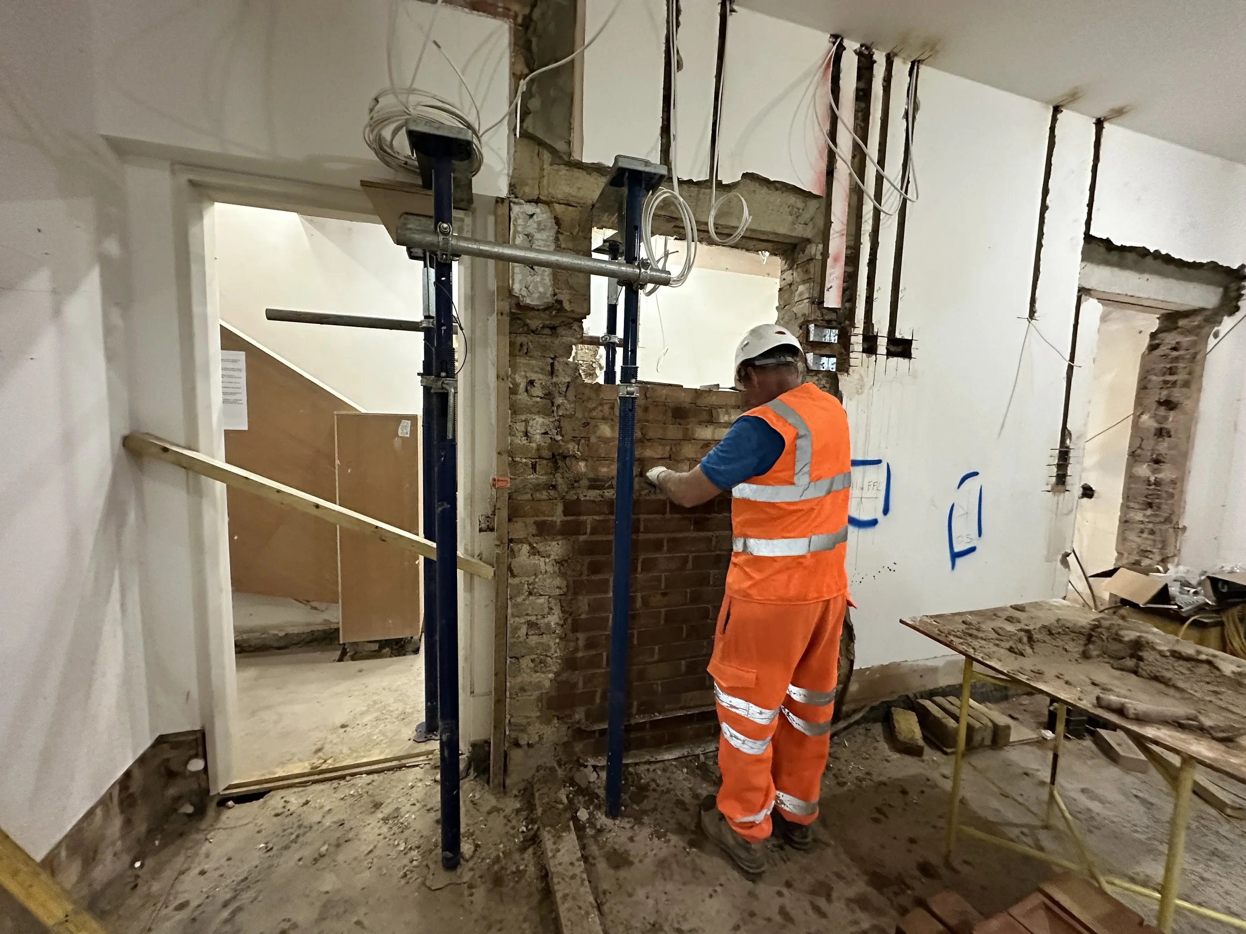 A construction worker wearing an orange safety vest, white helmet, and glasses working inside a building with exposed walls and electrical wiring, surrounded by construction materials and scaffolding.