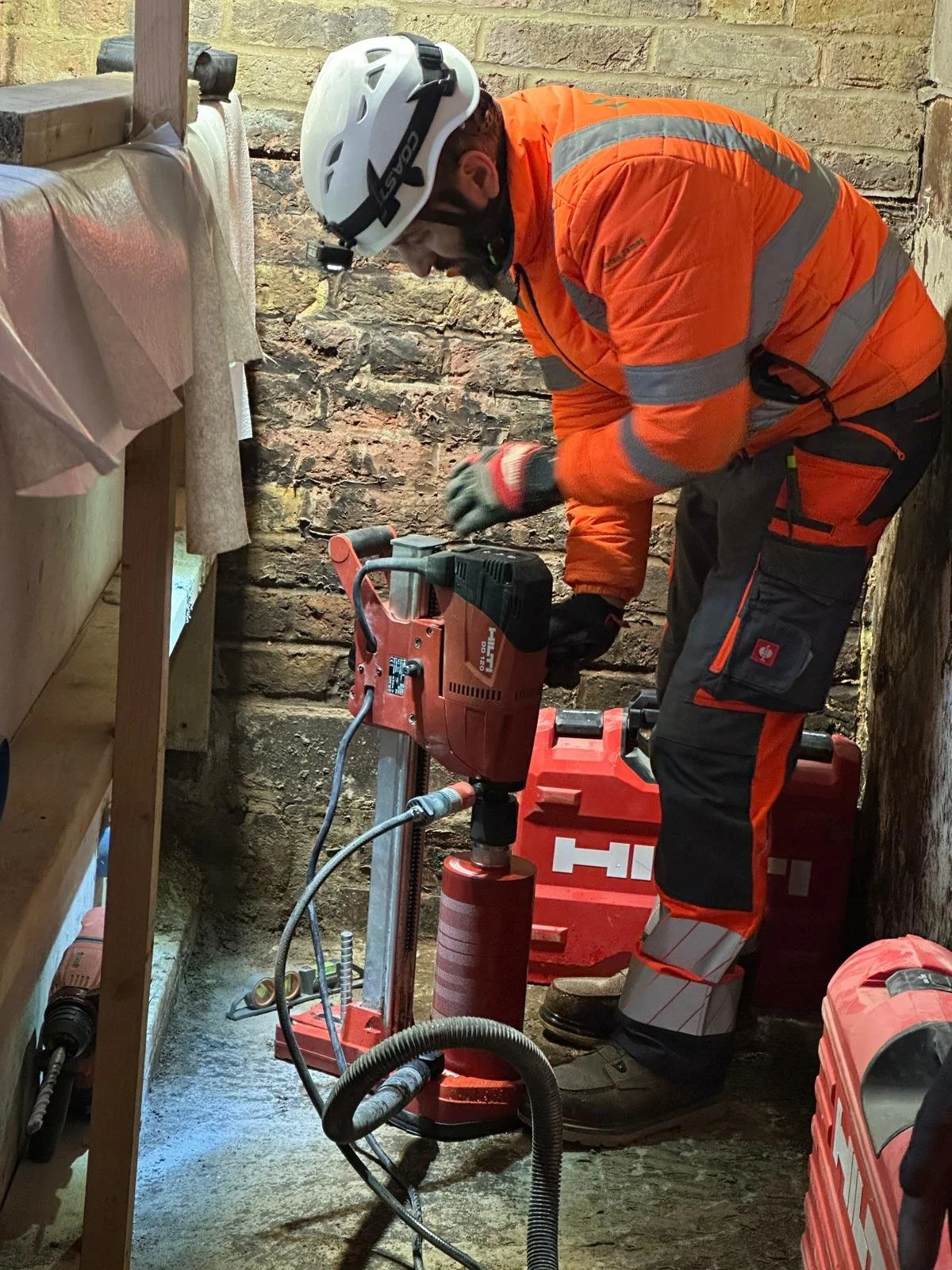 A worker in an orange safety jacket and helmet using a Hilti power tool to drill into a brick wall in a construction or renovation setting.