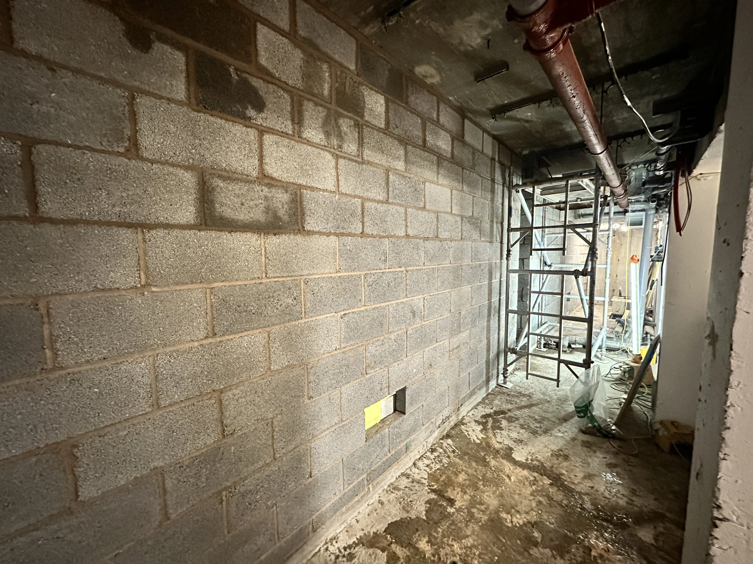 Construction site showing a brick wall with a hole, scaffolding, pipes, miscellaneous construction materials, and debris on the ground.