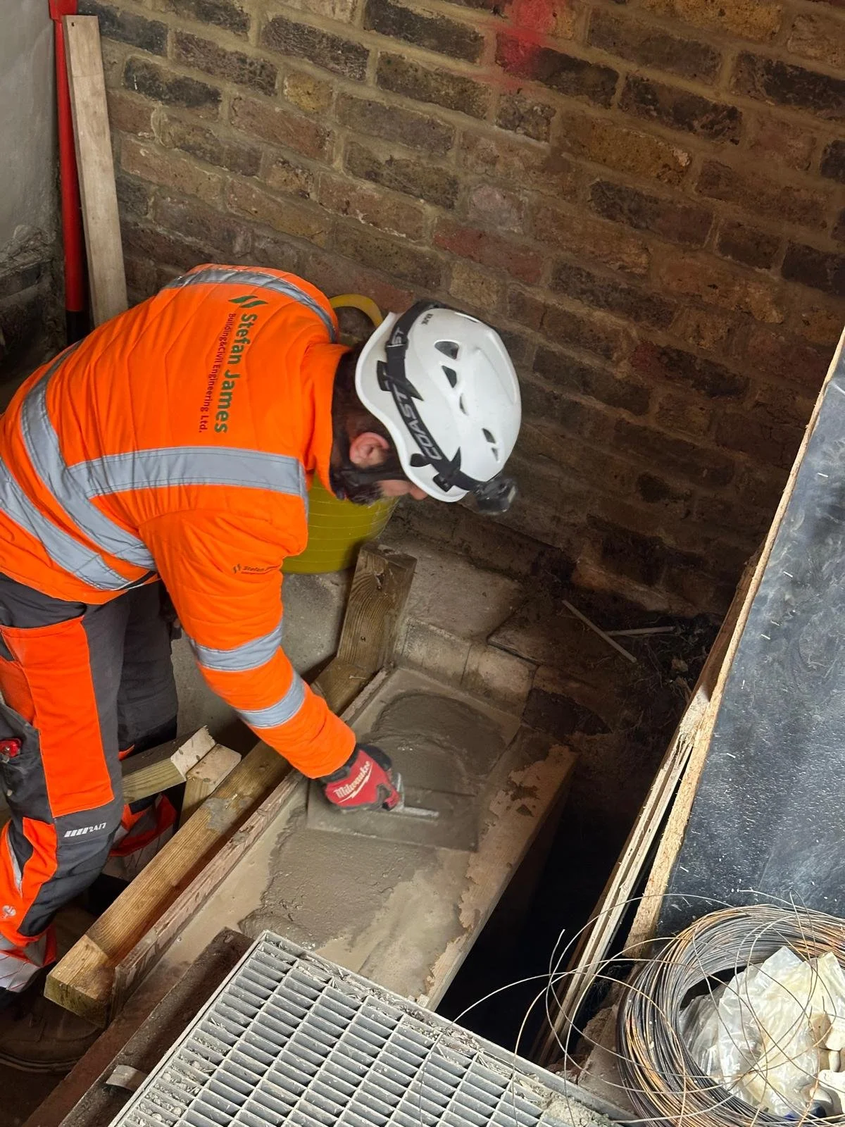 A construction worker wearing an orange safety vest, a white helmet, and gloves is pouring concrete into a form at a construction site. The worker is bending over with the back facing the camera. There is a brick wall behind him and construction tools around him.
