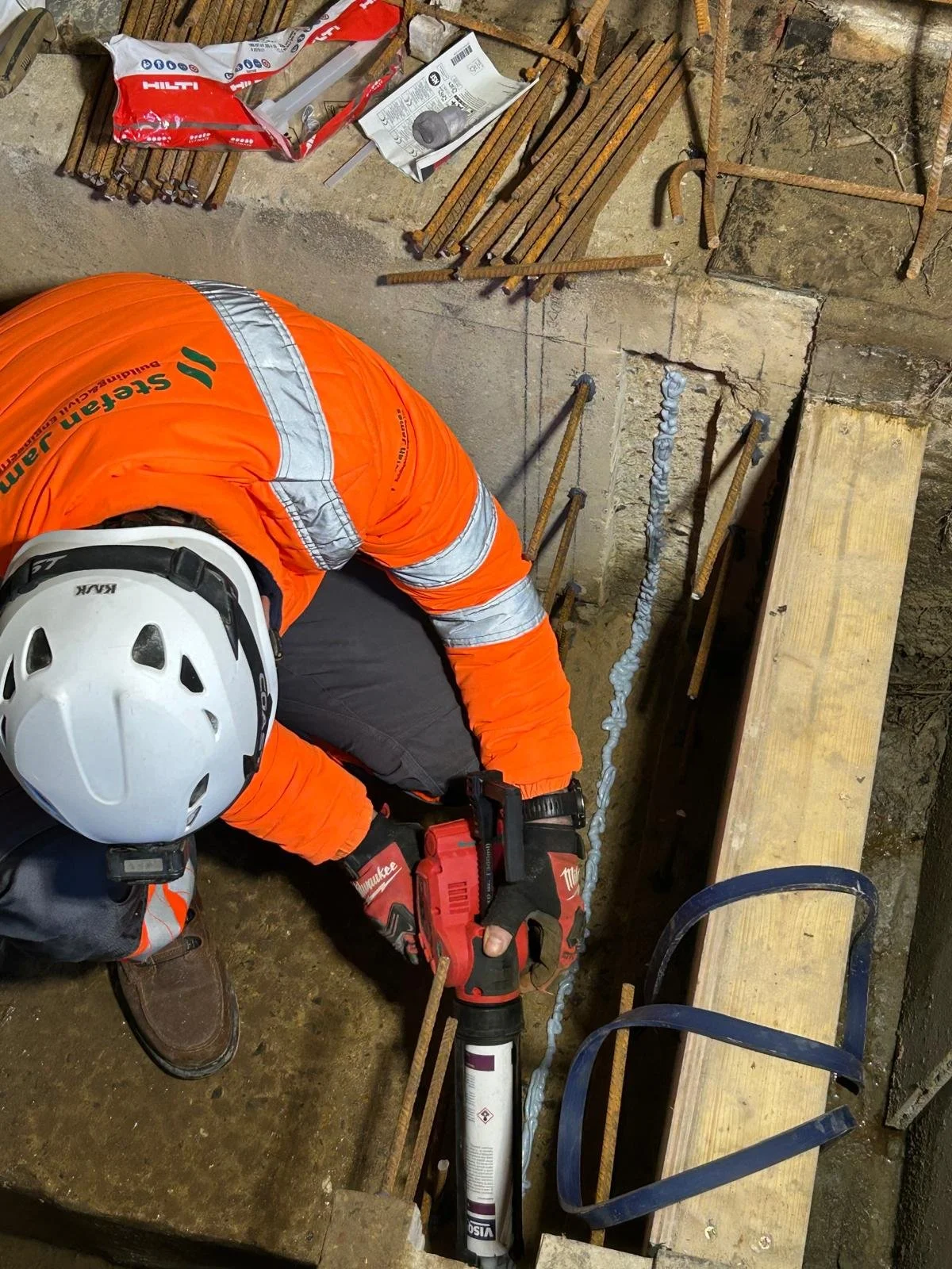 A construction worker wearing an orange safety jacket and a white helmet is working on a construction site, installing rebar in a concrete foundation or wall. The worker is using a power tool, likely a caulking gun or a similar device, to apply some material to the rebar. There are various pieces of rebar, tools, and construction materials scattered around the work area, including a bag of construction supplies and a wooden plank.