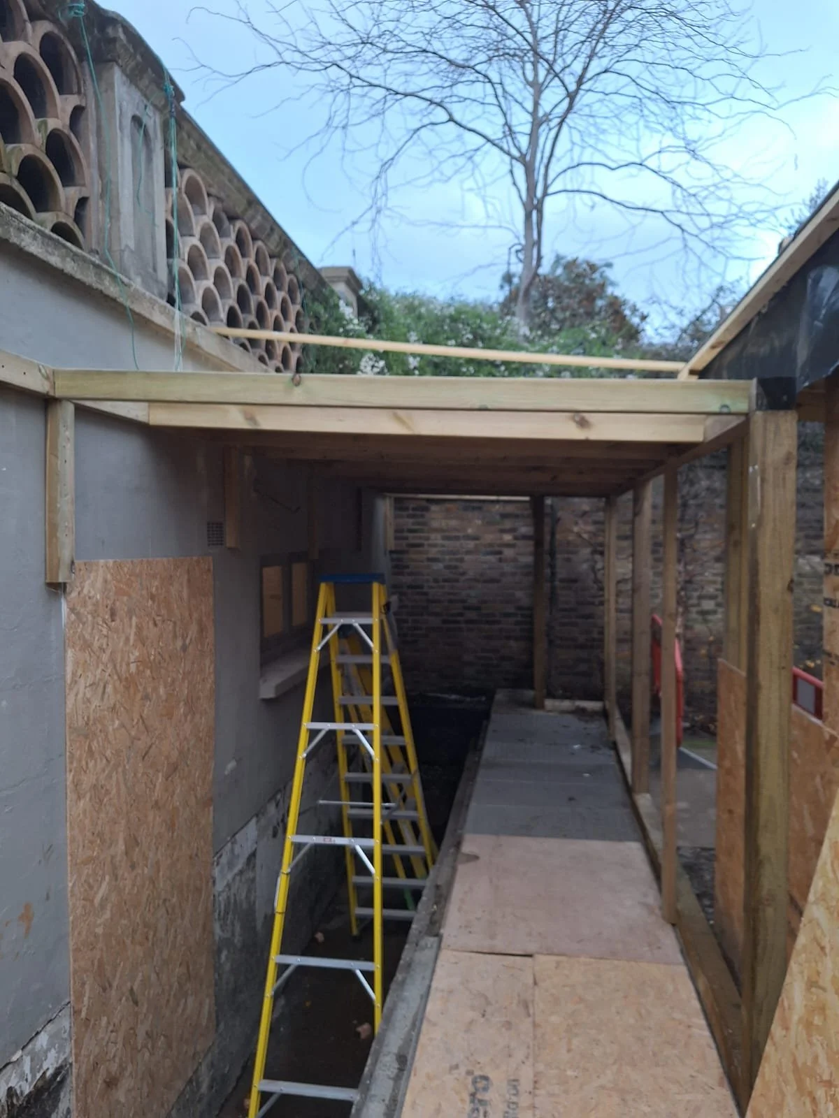 Construction site showing a wooden frame structure with a ladder inside, with plywood sheets on the ground and a partially built roof above.