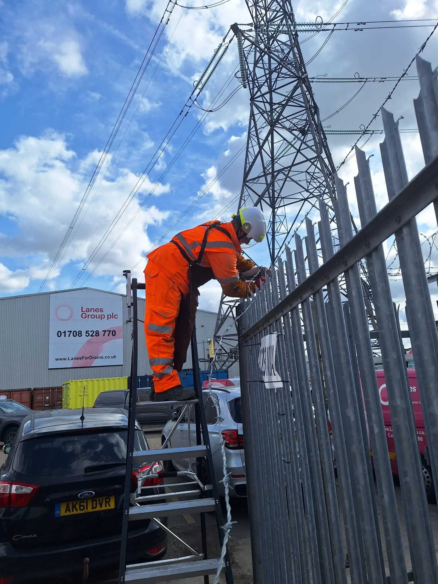 A worker in orange safety clothing and a white helmet is working on an electricity pole, using tools on a ladder. There are parked cars and industrial buildings in the background, under a partly cloudy sky.