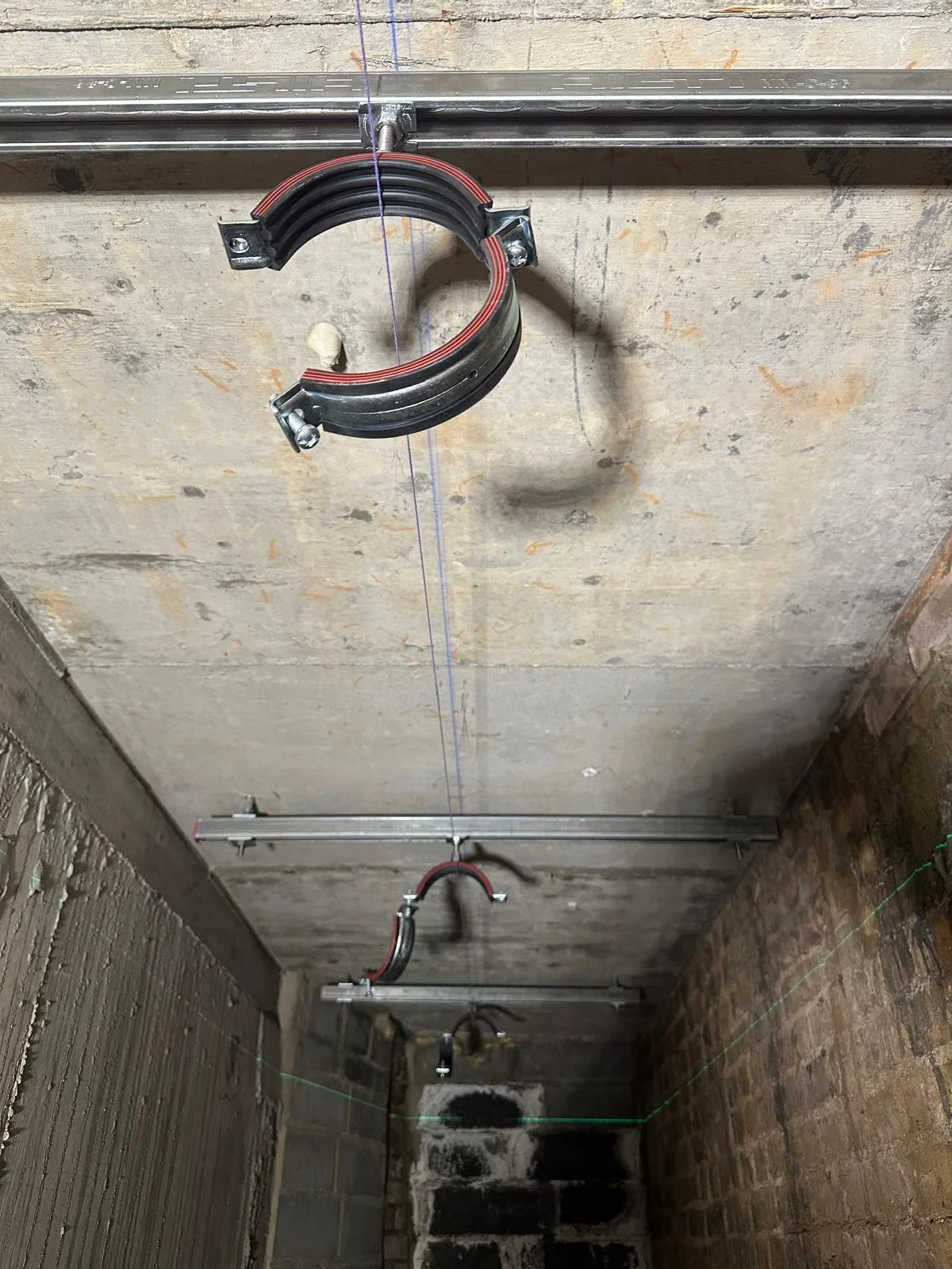 A construction site with metal pipes and a curved bracket mounted on a concrete ceiling, with green string lines marked at various points.
