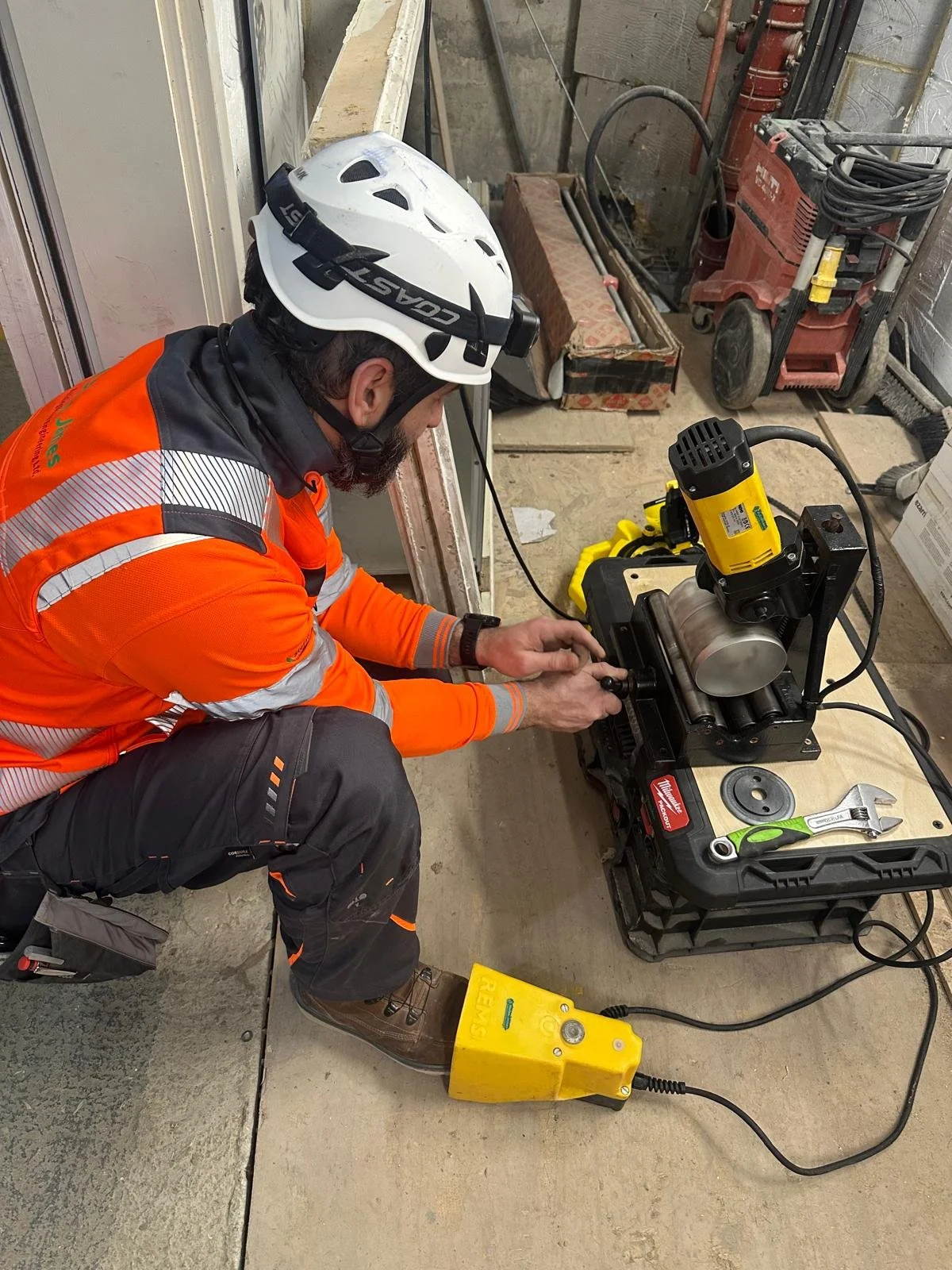 A construction worker wearing an orange safety jacket, a white helmet, and brown boots is kneeling on the floor working with a metal roller machine connected to a yellow corded power supply at a construction site.