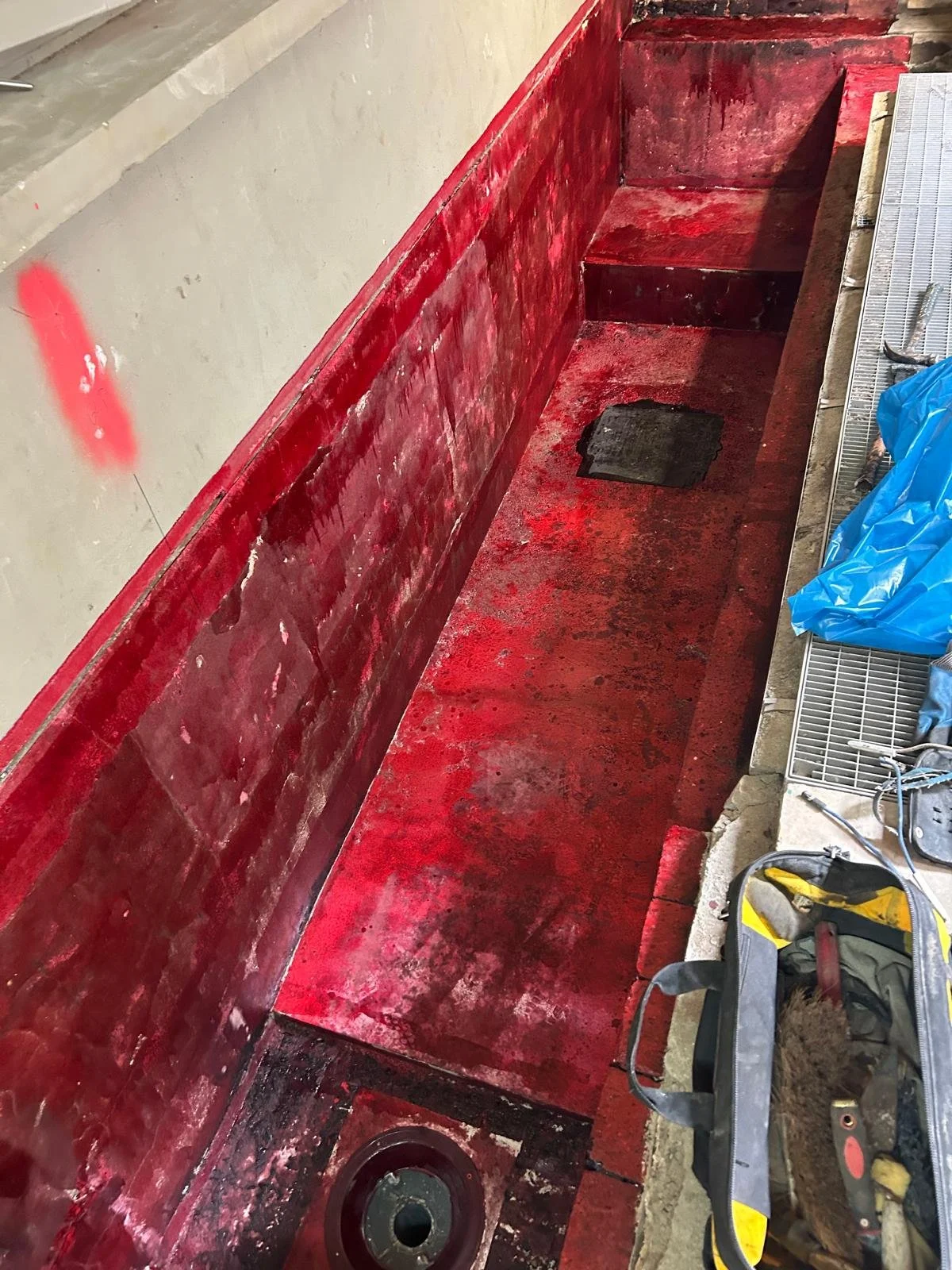 A section of a construction site with a red-painted area, possibly a bench or ledge, and construction tools like a level, screwdriver, and blue plastic bag nearby. The area appears to be in the process of renovation or repair.