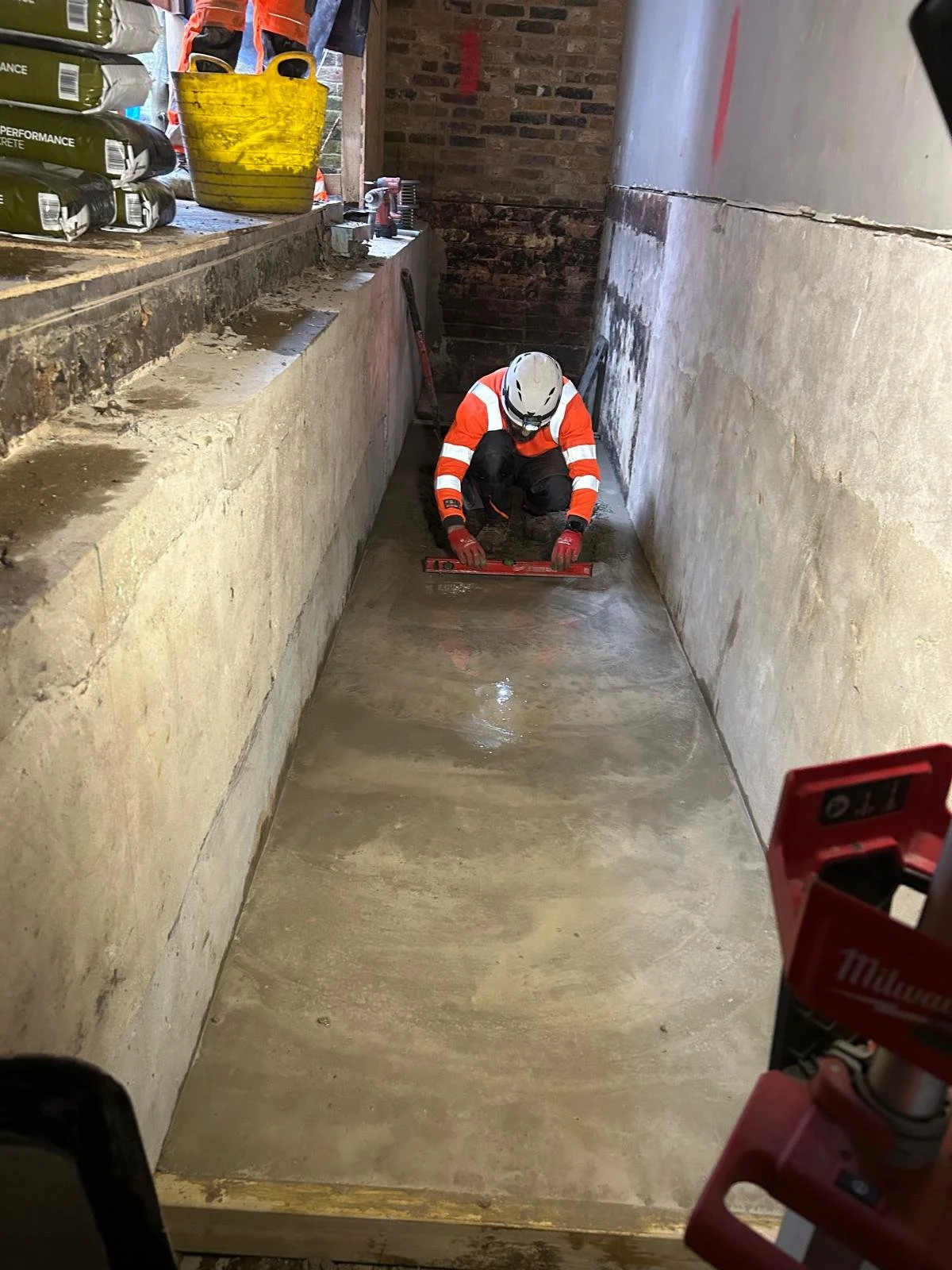 A construction worker wearing a white safety helmet, red gloves, and a high-visibility orange jacket kneels on the ground, using a level to check the surface in a narrow, partially finished concrete space. Construction tools, materials, and a yellow bucket are visible on a ledge to the left.