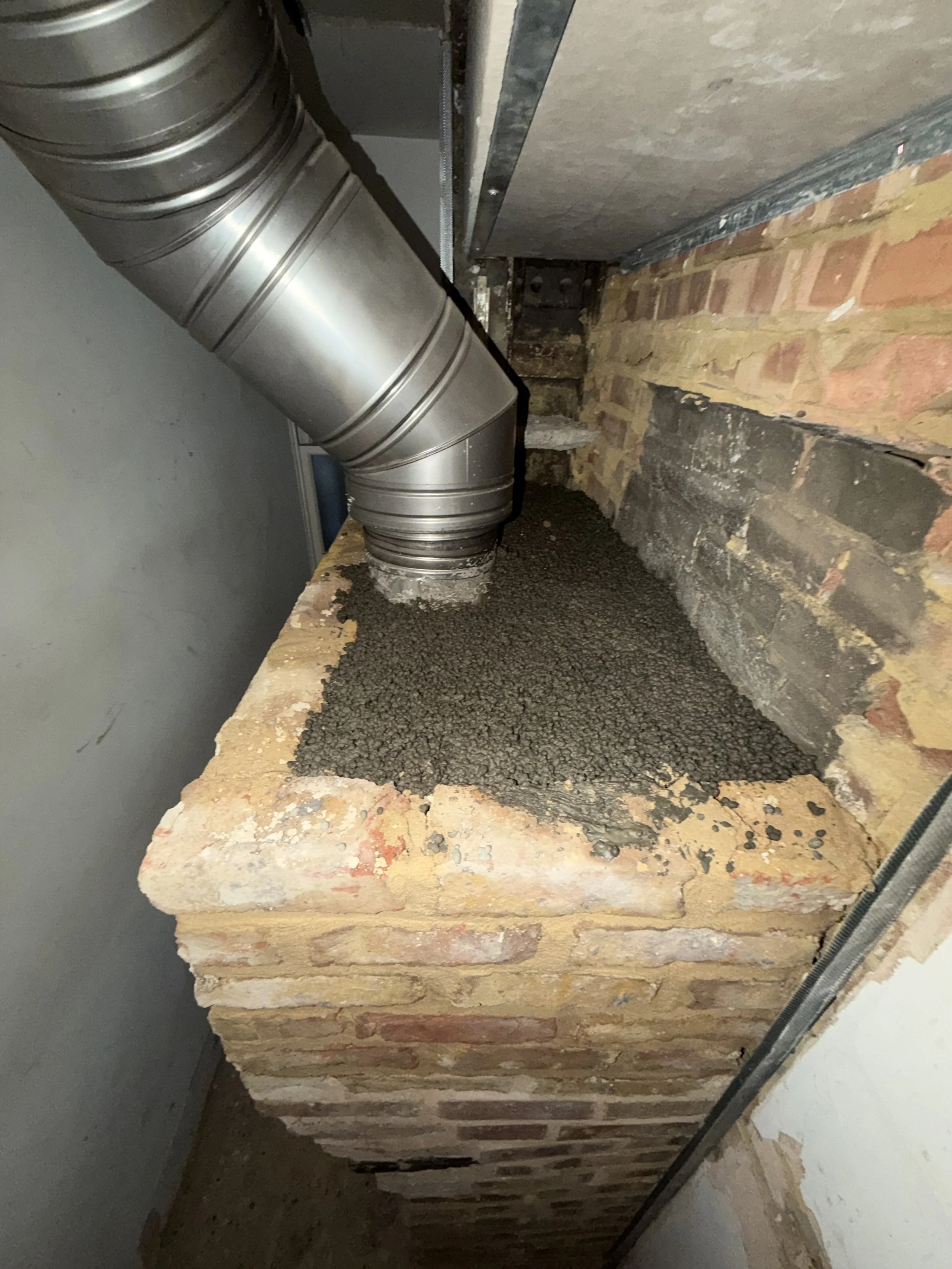 Construction site showing a brick wall with gravel spread on a wooden surface, a metal ventilation duct on the upper left, and construction tools or materials in the background.
