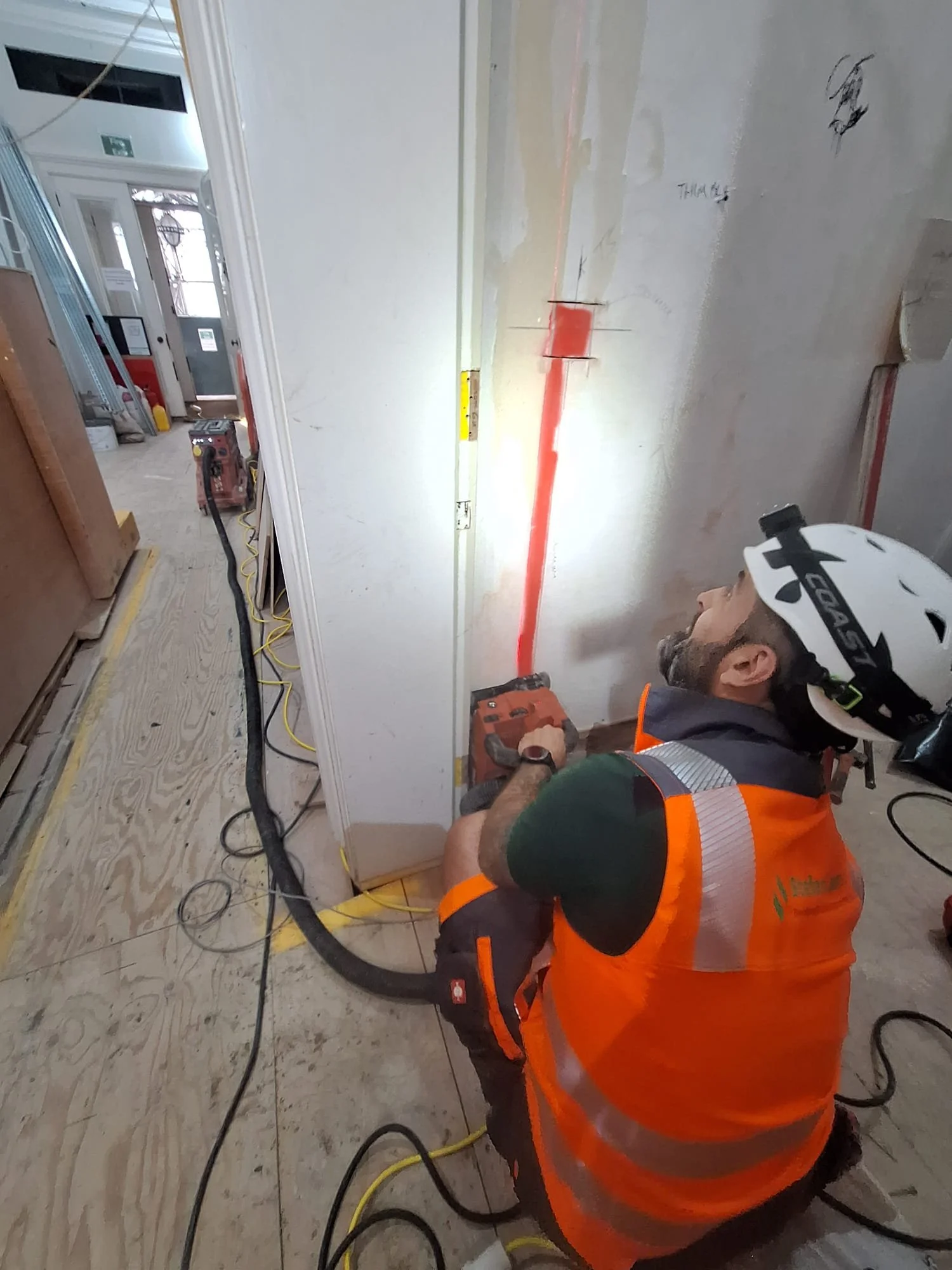 A construction worker wearing an orange safety vest and a white helmet working on an electrical or plumbing installation inside a building. The worker is kneeling and using tools near a wall with some markings and a red line. The area has various construction materials and tools.
