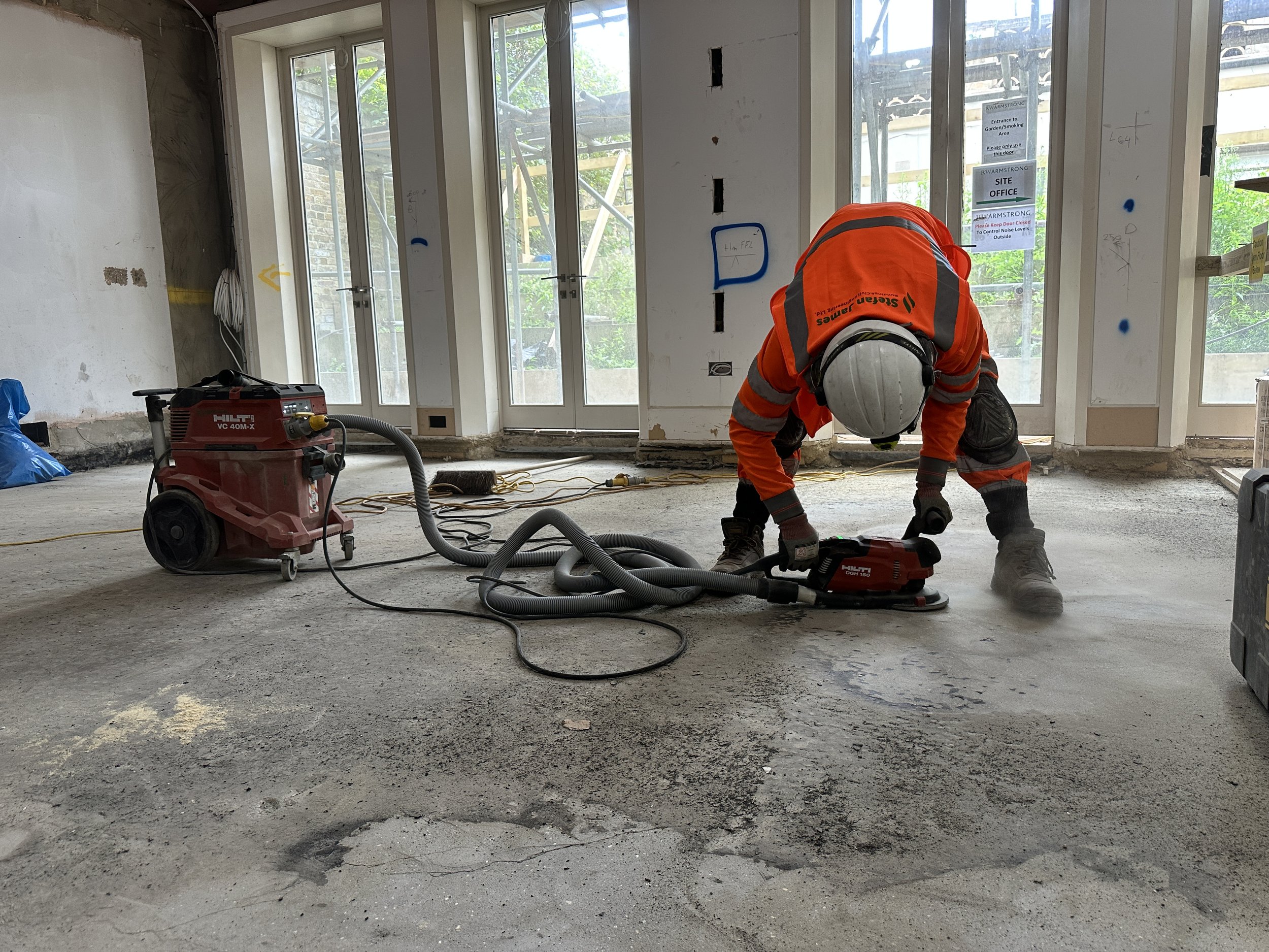 Construction worker in orange safety gear and helmet kneeling on concrete floor inside a room under construction, with large windows, construction tools, and scaffolding outside.