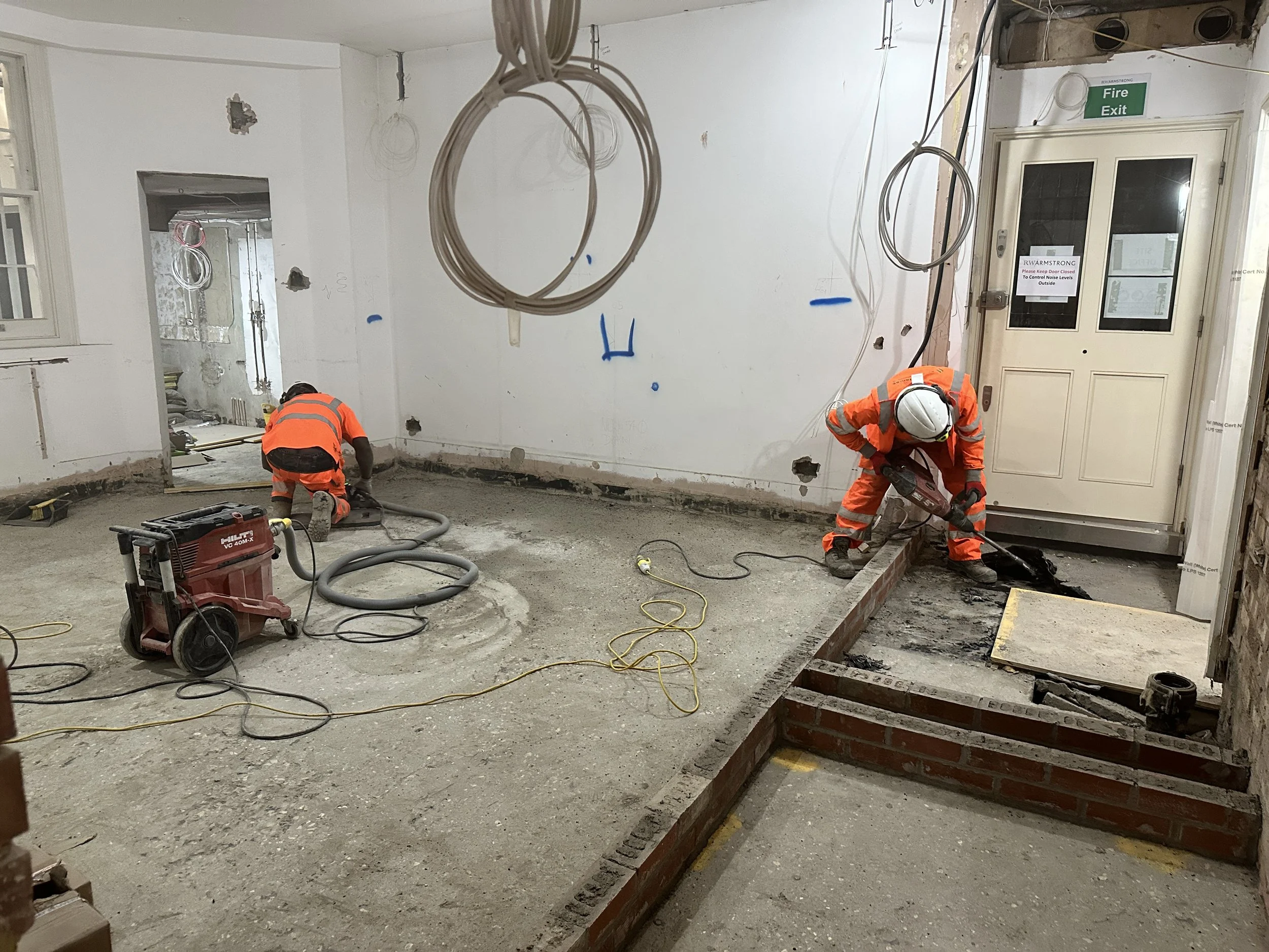 Construction workers in orange safety gear working on the floor of a building under renovation, with exposed wiring and partially finished walls.