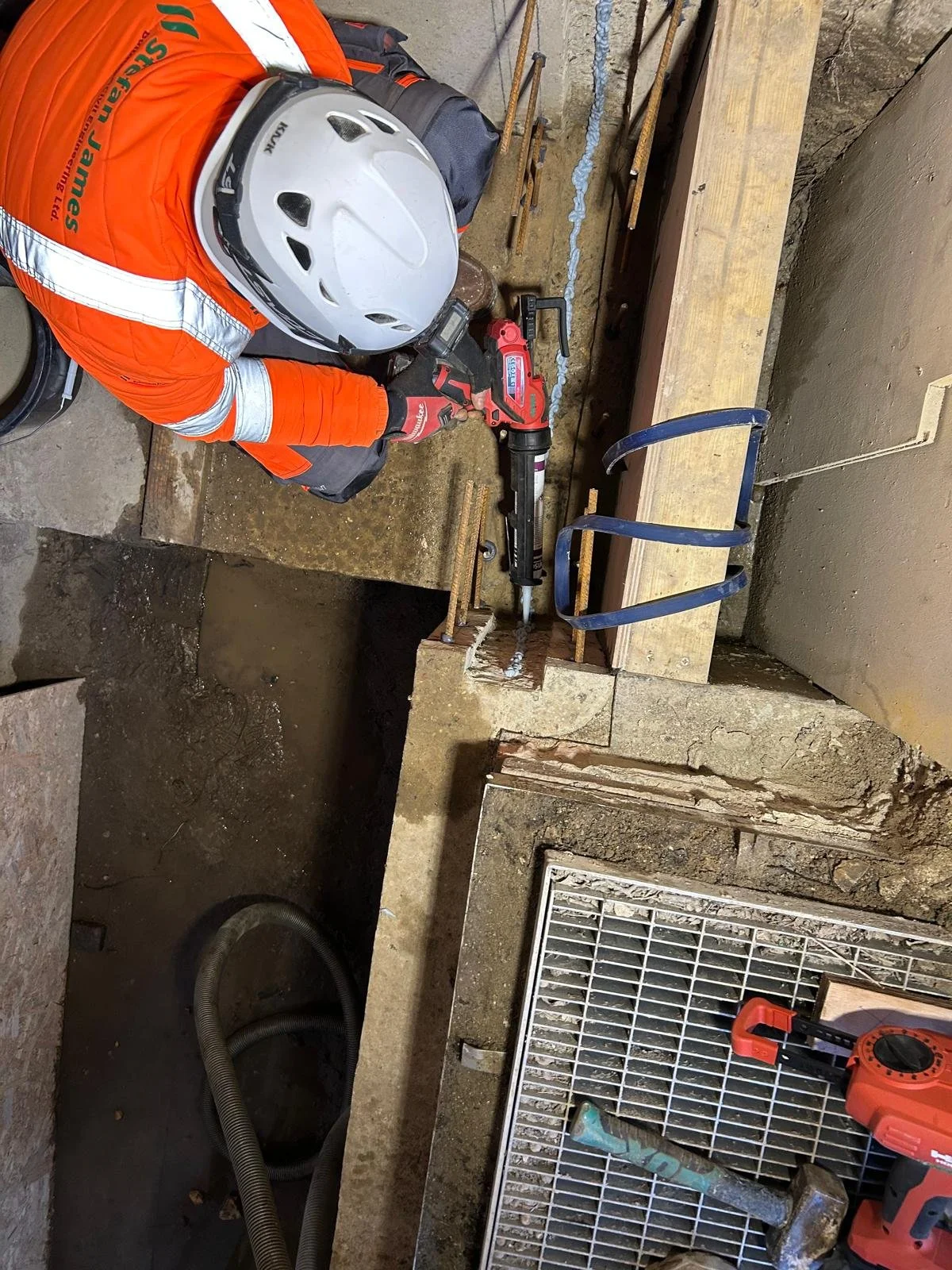 Worker in an orange safety jacket and white helmet using a power tool on a construction site with wooden and metal debris, hoses, and building materials.